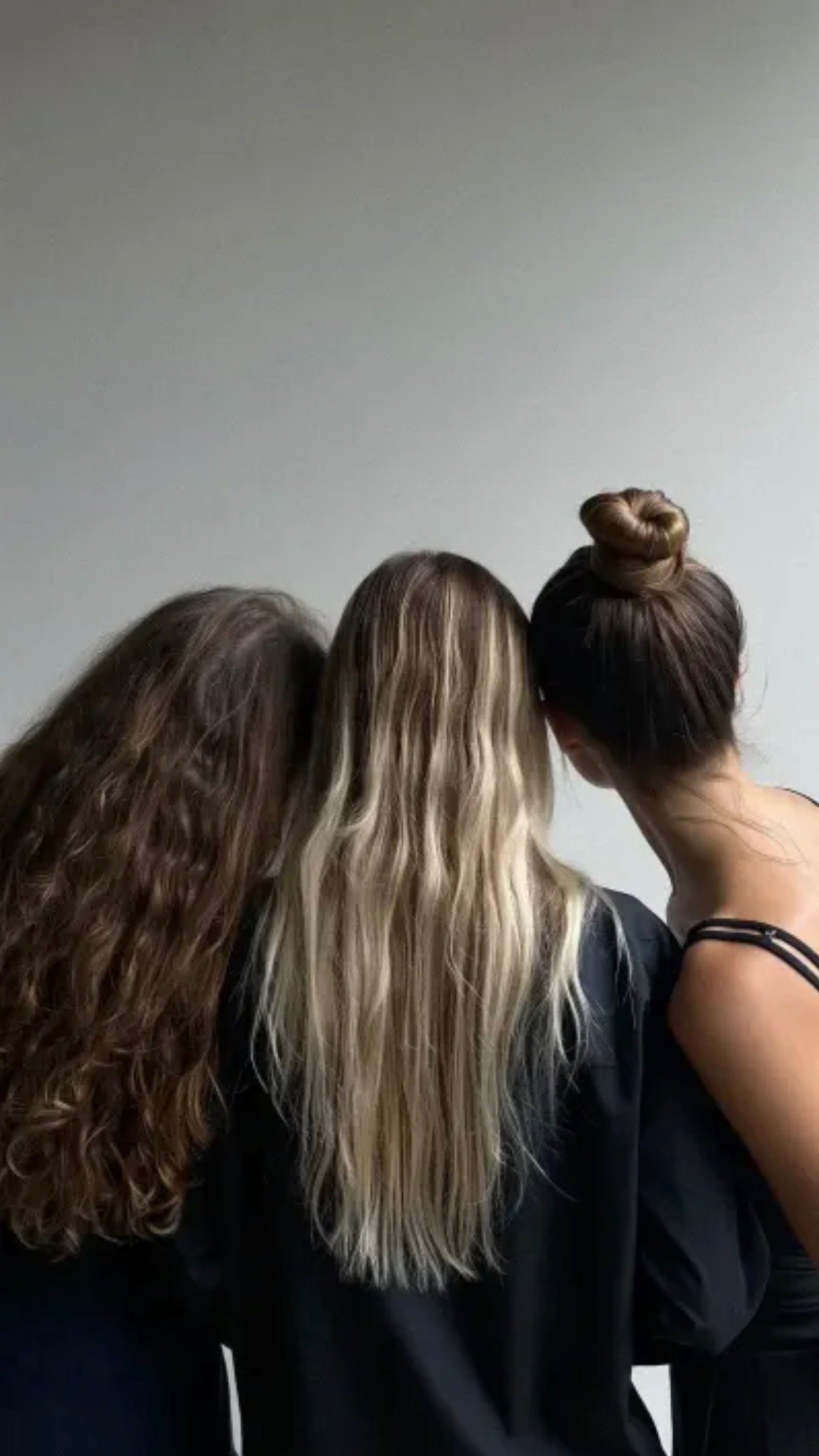 Three women with different hairstyles leaning their heads together, showing the back of their heads against a plain white wall.