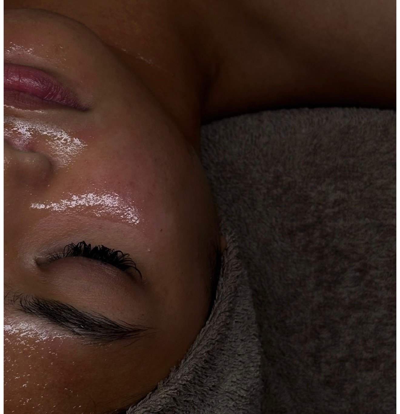 Close-up of a woman's face tilted sideways, showing closed eyes and shiny skin, with a gray fabric covering part of her face.