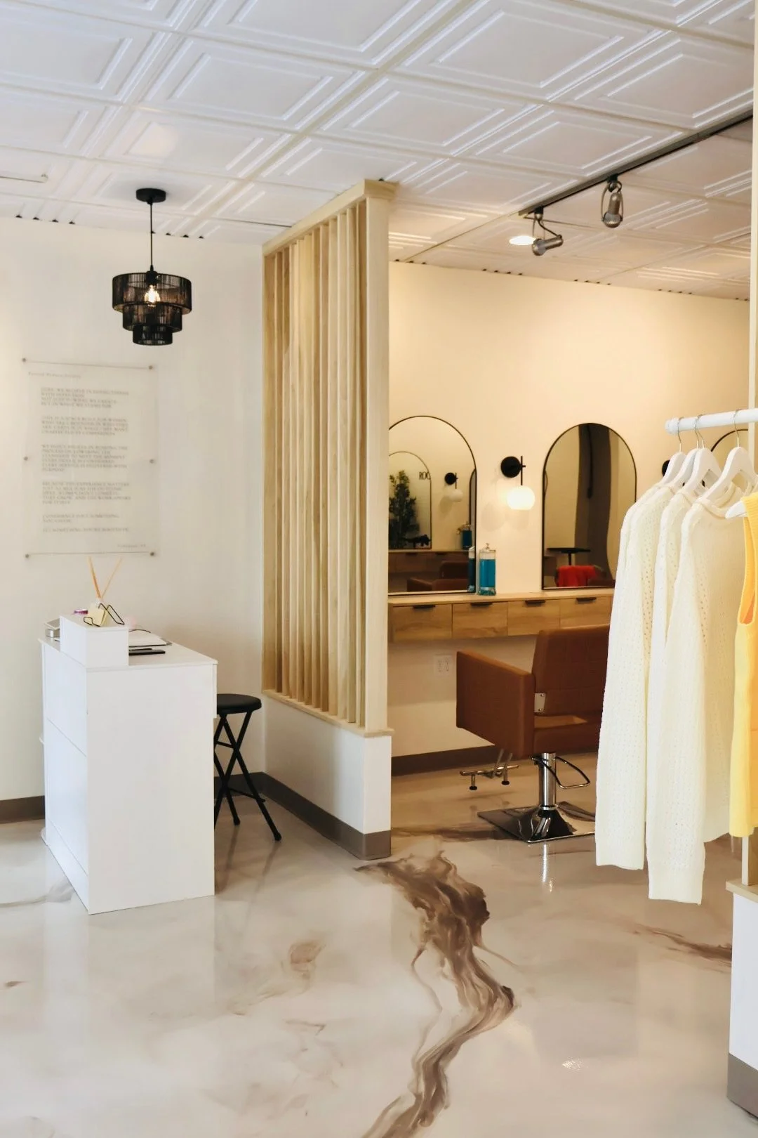 Interior of a modern hair salon with a reception area, wooden divider, mirrors, styling chairs, and hanging clothes in yellow and white.