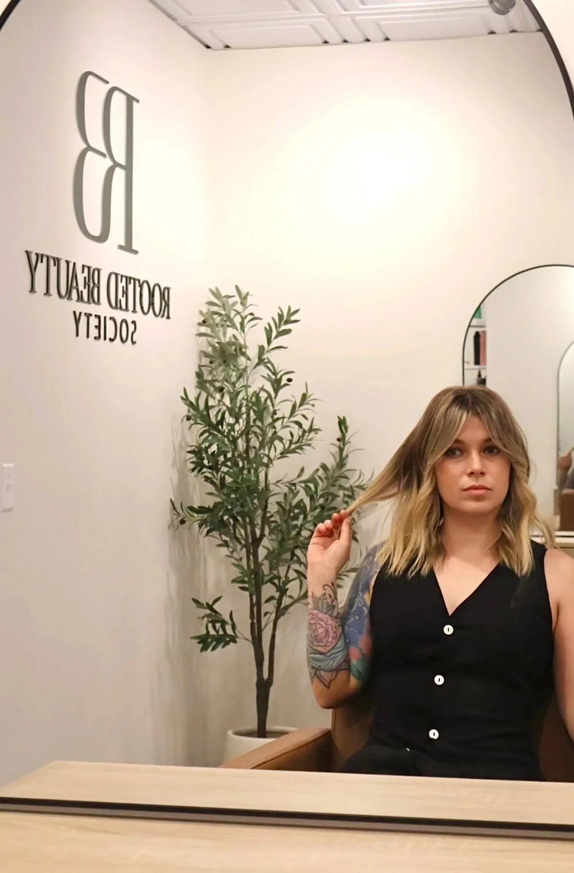 A woman with blonde, wavy hair and tattoos on her arm sitting in front of a mirror at a hair salon, with a potted tree and a sign on the wall that reads 'ROOTTED BEAUTY SOCIETY.'