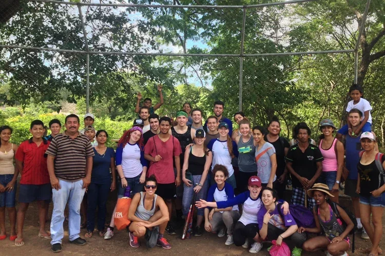 Group of diverse people gathered on a baseball field surrounded by trees, smiling and posing for a photo.