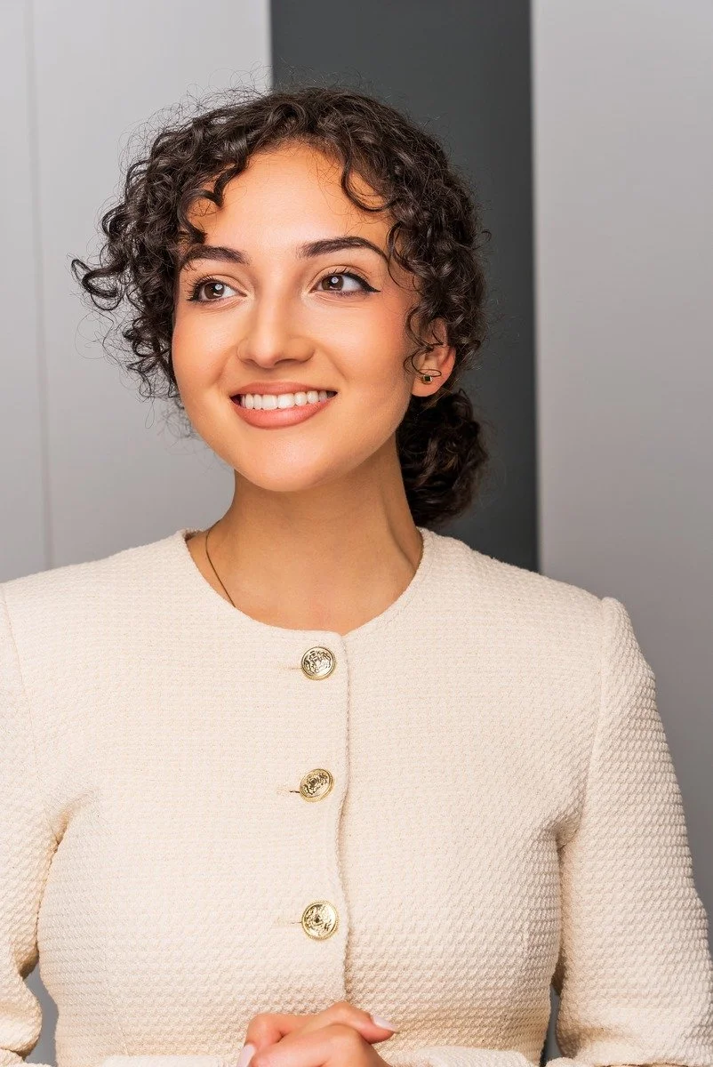 A young woman with curly brown hair, smiling, wearing a cream-colored textured jacket with gold buttons, standing indoors against a gray background.
