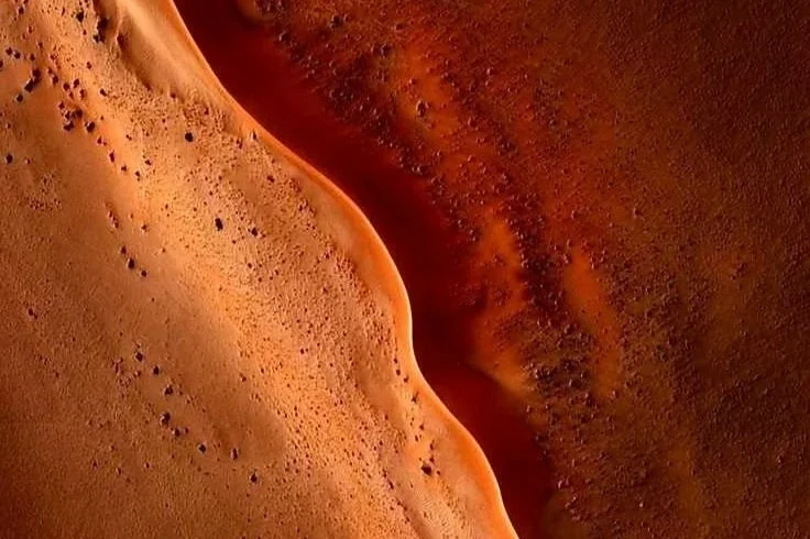 Close-up of a red-brown sandy desert landscape with a wavy pattern and small scattered rocks or pebbles.