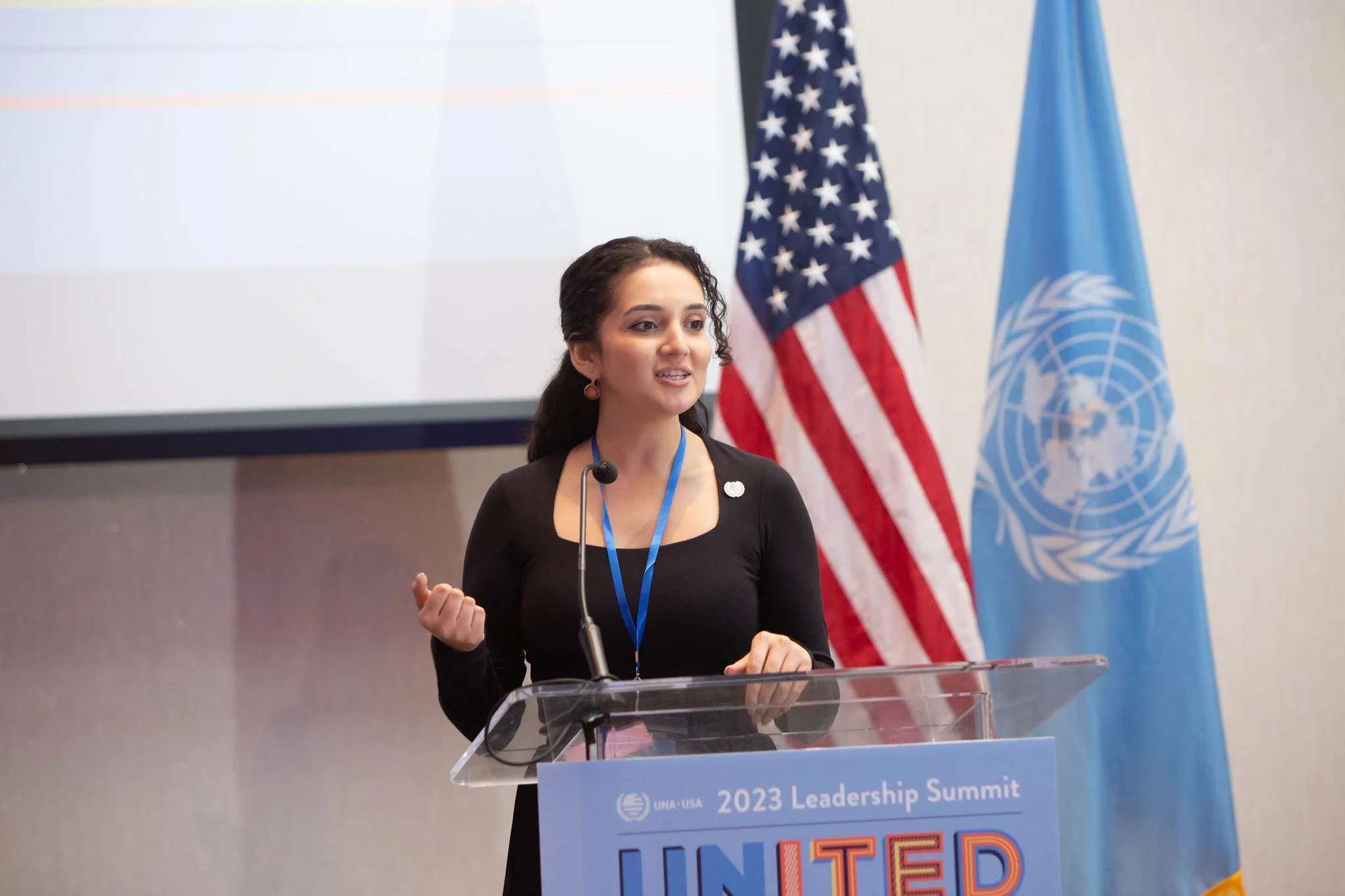 A young woman speaking at a podium during the 2023 Leadership Summit, with the United Nations flag and the U.S. flag behind her.