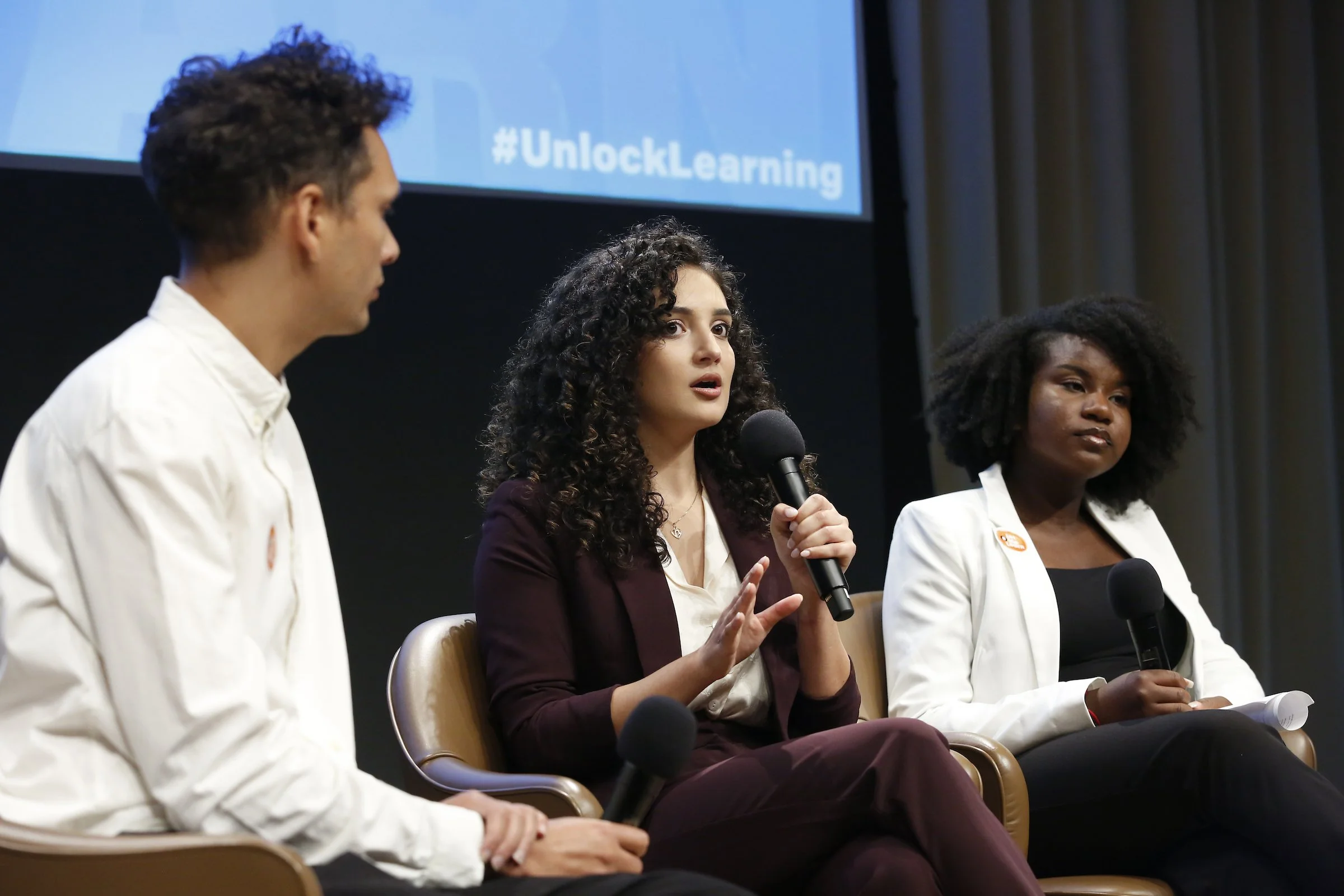 Three individuals sitting on a stage during a panel discussion, one woman holding a microphone and speaking, with a projection screen behind them displaying the hashtag #UnlockLearning.