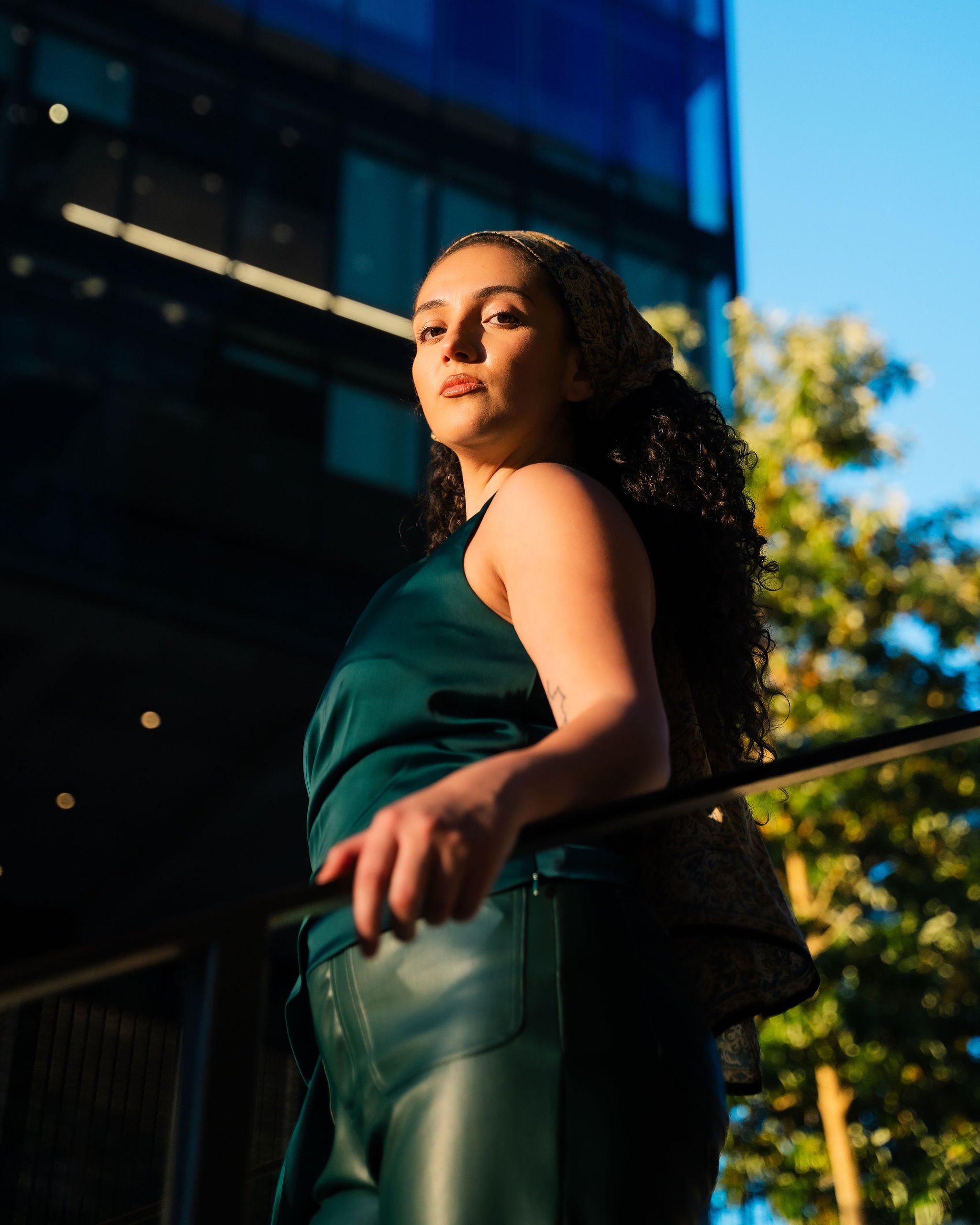 A woman with curly dark hair wearing a green sleeveless top and matching green pants, standing outdoors next to a metal railing, with modern glass buildings and trees in the background during daytime.