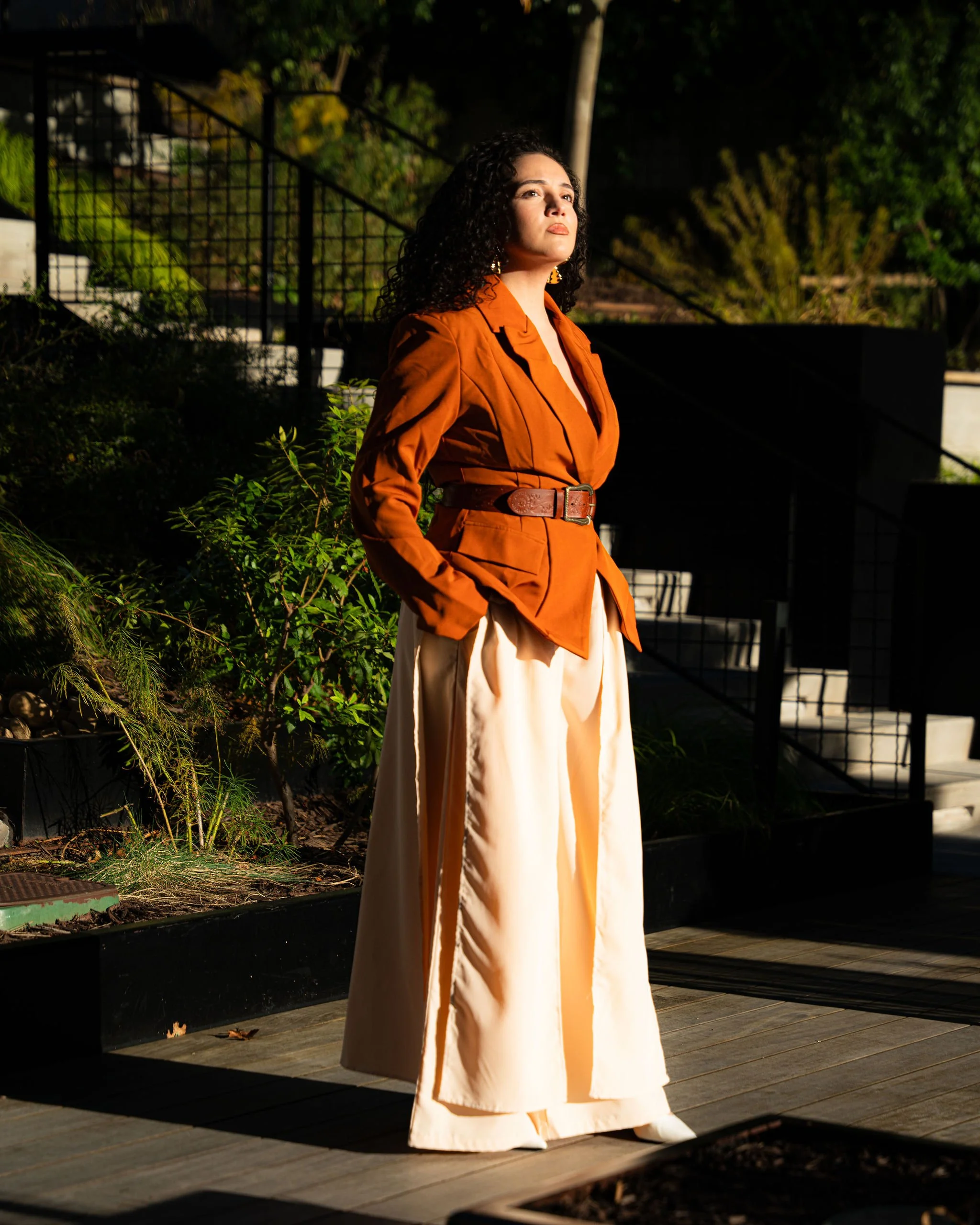 A woman with dark curly hair standing outdoors at night, wearing a rust-colored blazer and high-waisted cream pants, illuminated by warm sunlight.