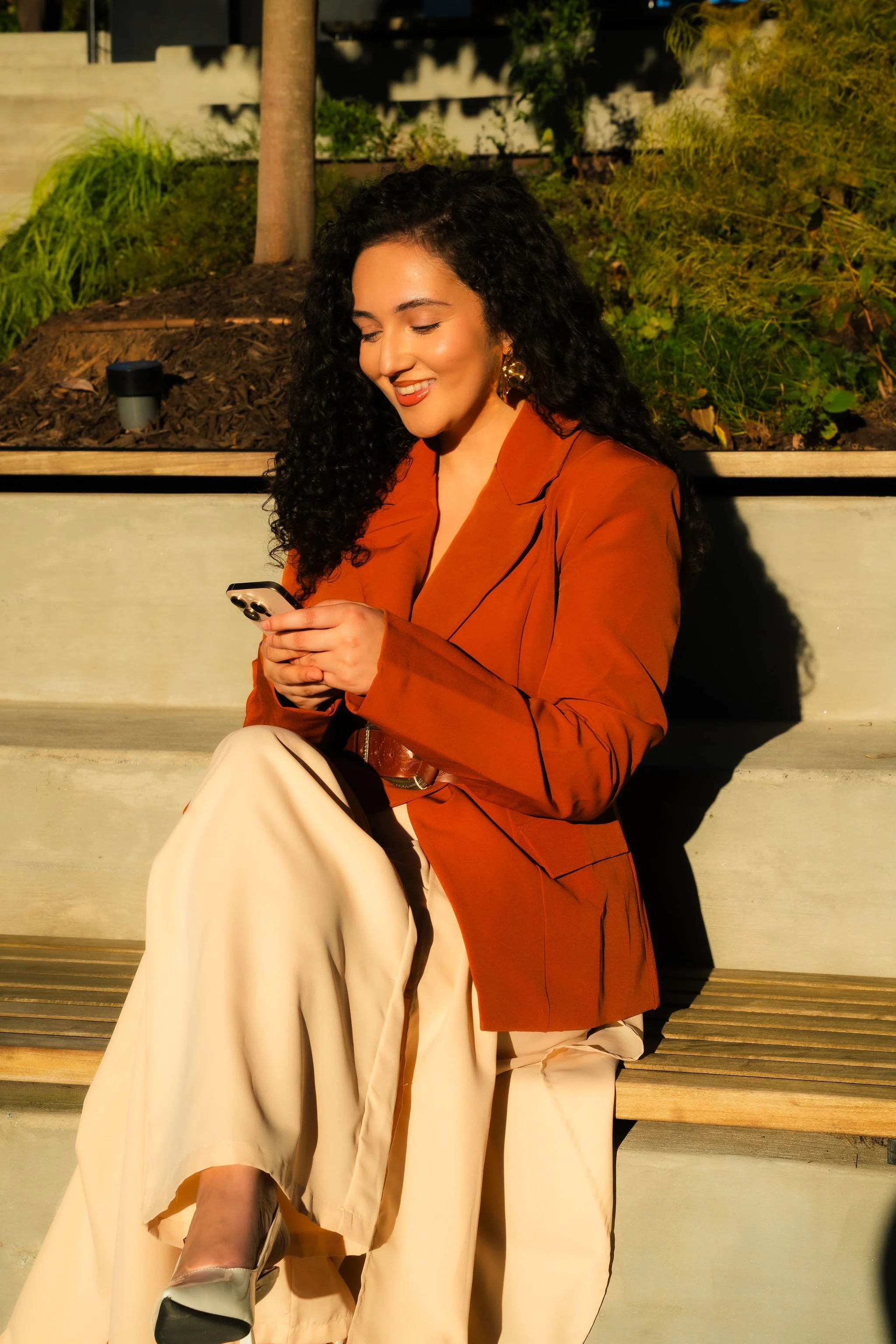 A woman with curly black hair smiling and looking at her phone while sitting on a bench outdoors, wearing a rust-colored blazer and cream-colored wide-leg pants.