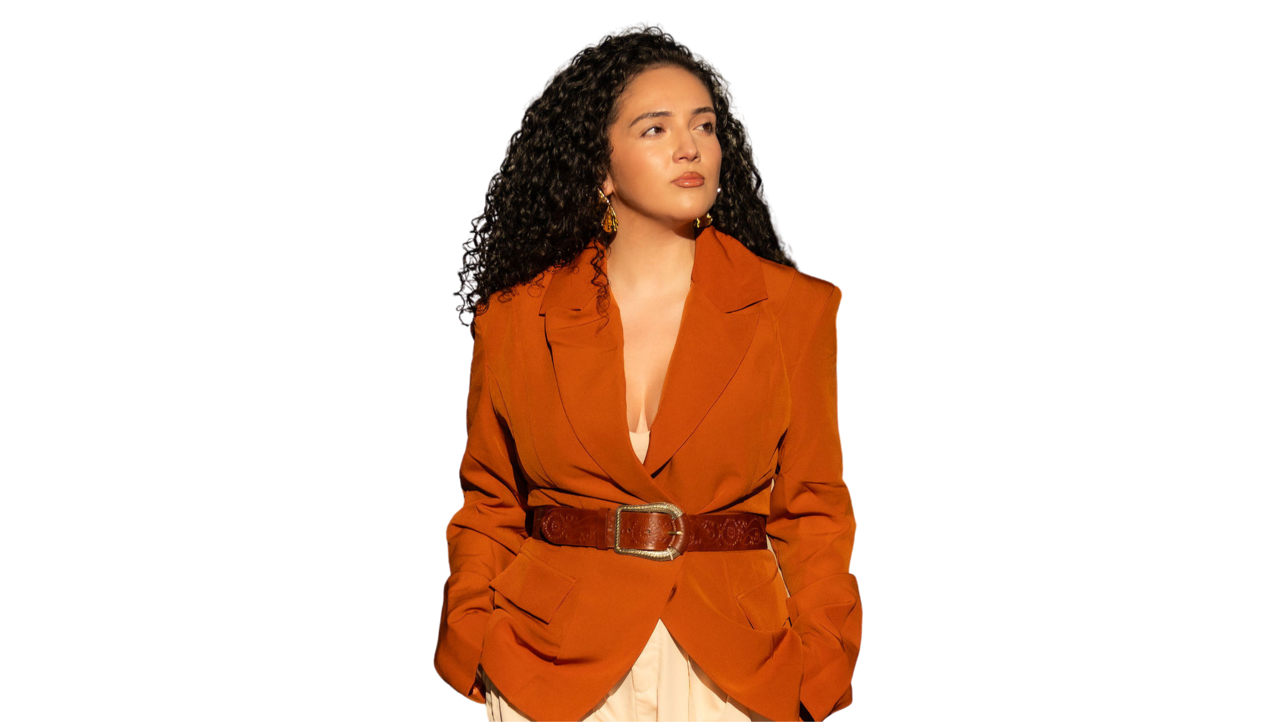 A young woman with curly dark hair and light skin, wearing a rust-colored blazer with a matching belt, white dress, and gold earrings, looks to the side.