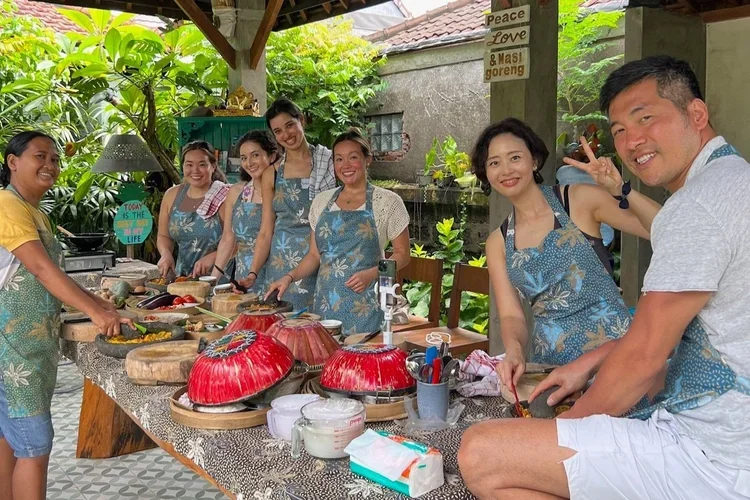 Group of seven people, mostly women, wearing aprons and cooking together outdoors under a pavilion, with a man sitting at the end of the table making a peace sign. The table is filled with cooking ingredients and utensils, with tropical plants in the background.