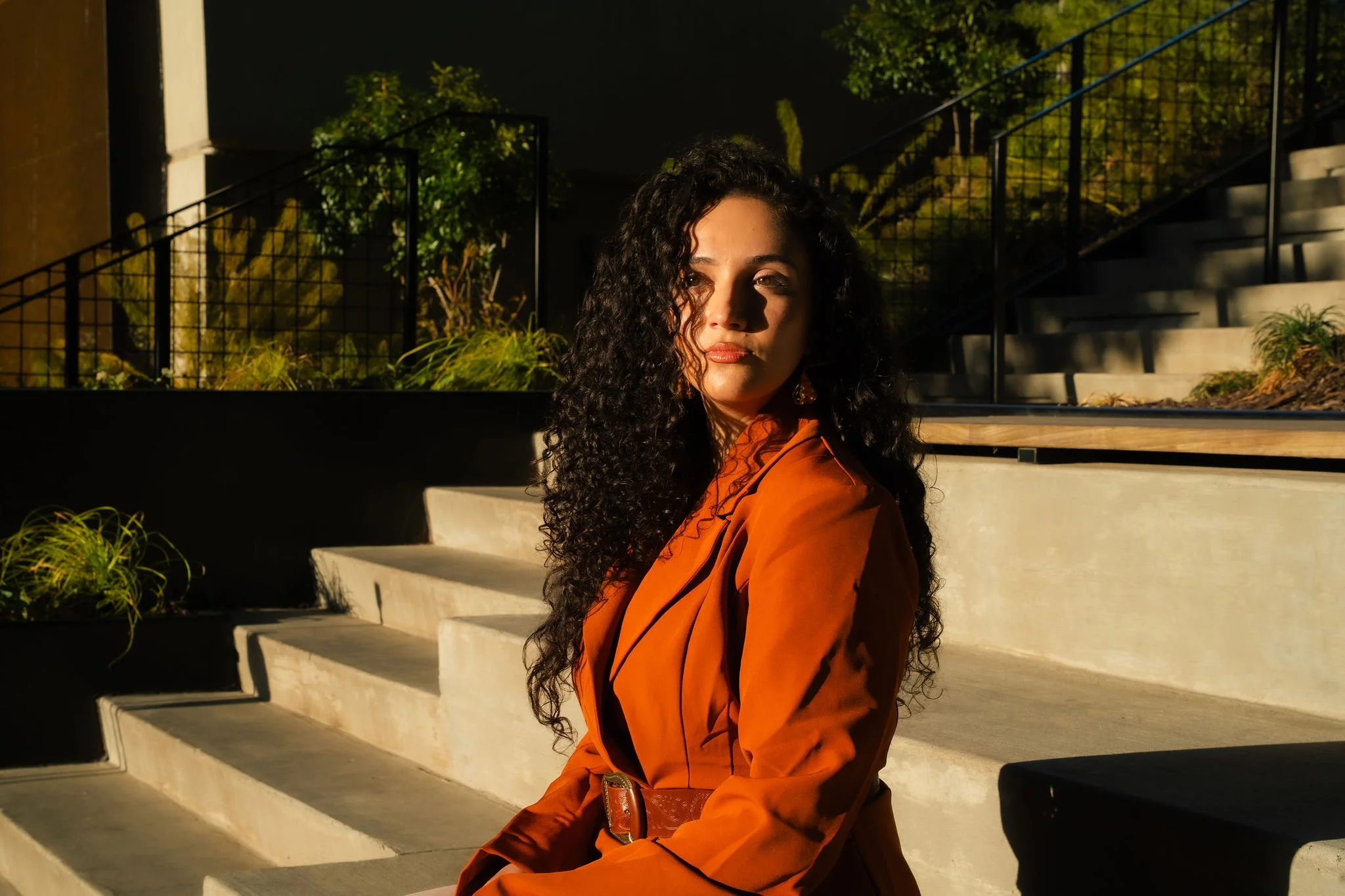 A woman with curly dark hair wearing an orange coat stands on an outdoor staircase at sunset. She looks over her shoulder with a serious expression, with greenery and modern black railings in the background.