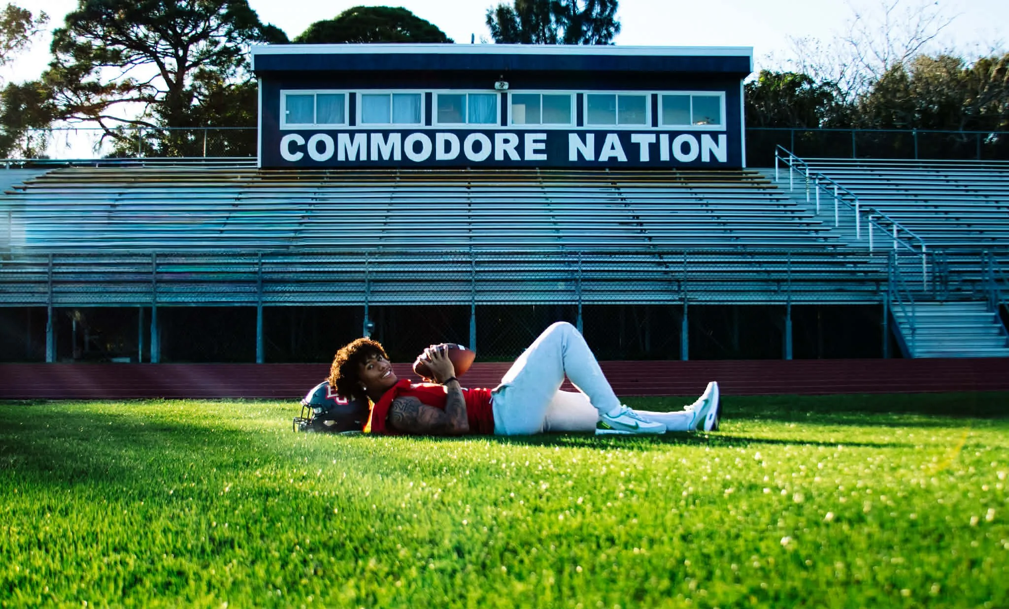 A person lying on the grass football field holding a football, with empty bleachers and a sign reading 'COMMODORE NATION' in the background.