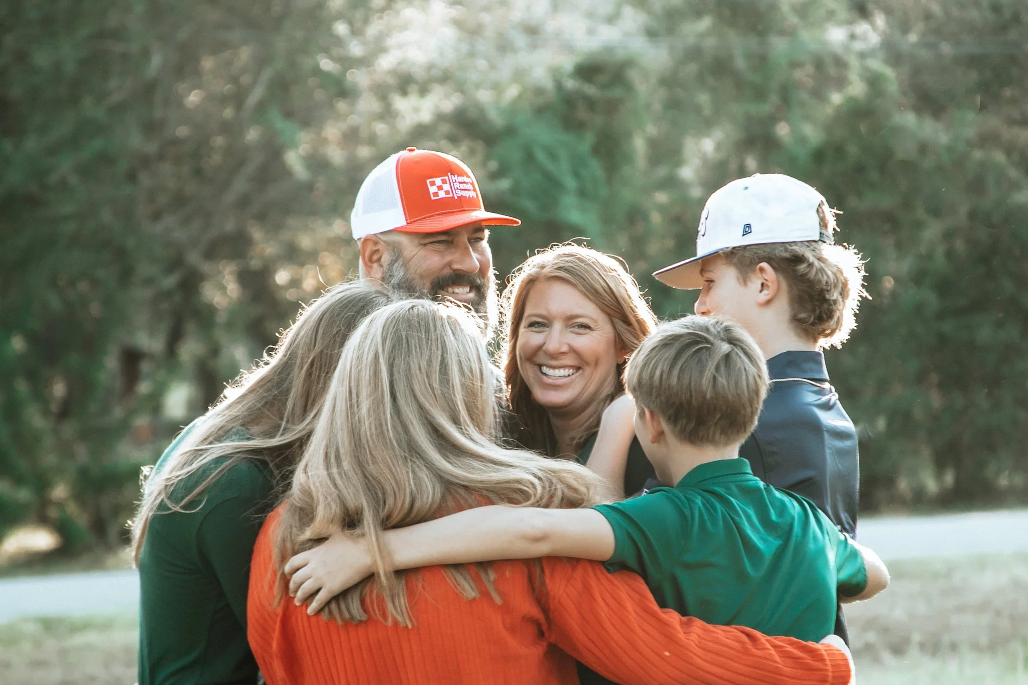 A group of six people, including two adults and four children, are hugging outdoors in a park with trees in the background. They're smiling and appear happy.