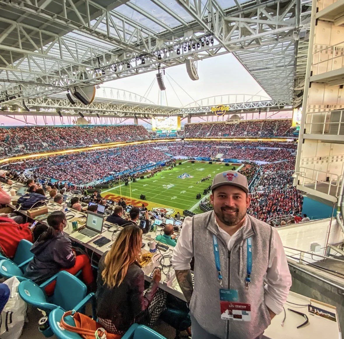 A man smiling at the camera inside a stadium during a football game, with a large crowd in the stands and the field visible in the background.