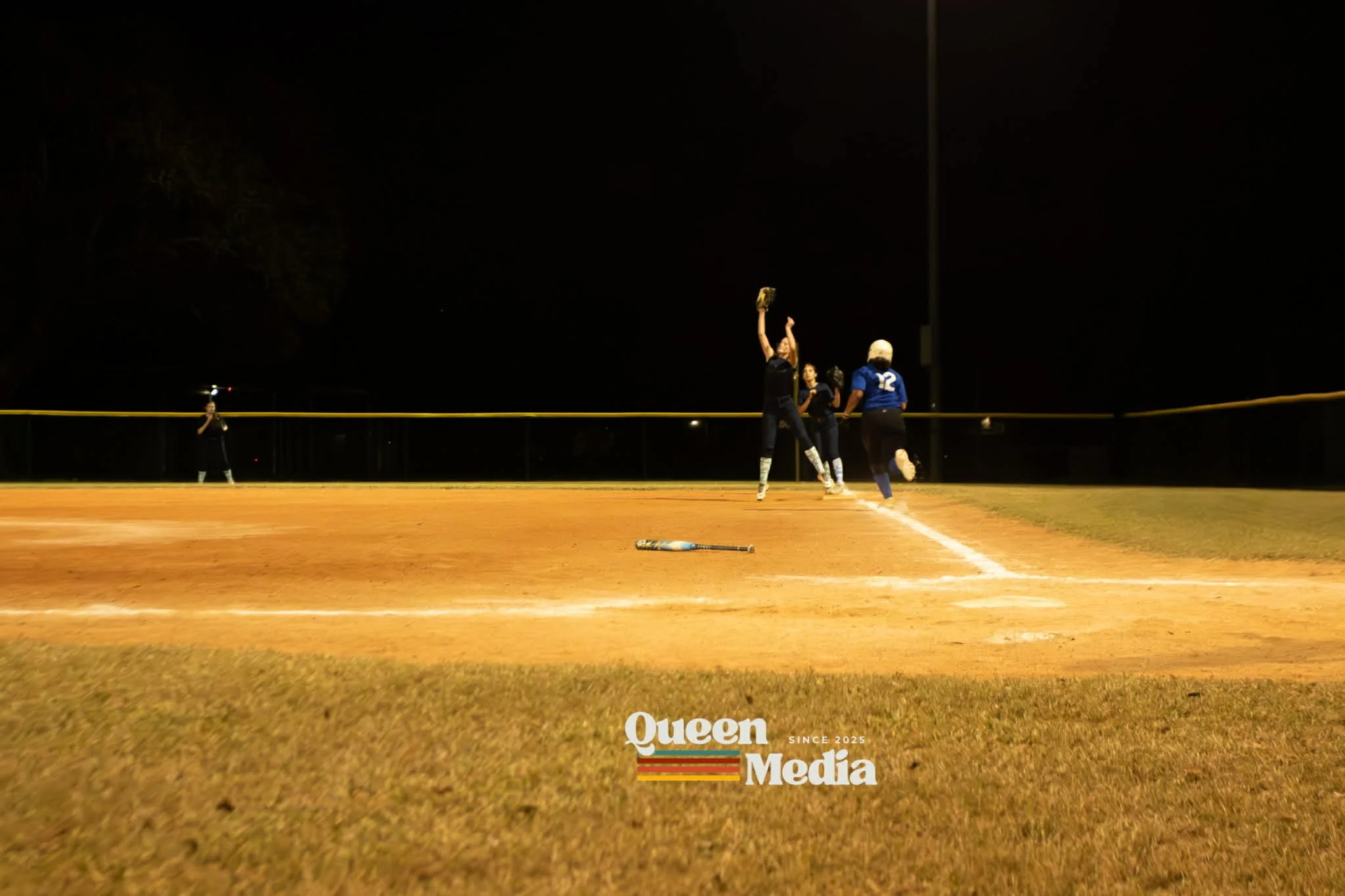 Softball players celebrating on a field at night with a dark sky, and a fence in the background. One player jumps with a glove raised, others run toward home plate, a bat lies on the ground, and an umpire stands near the fence. Queen Media logo at the bottom.