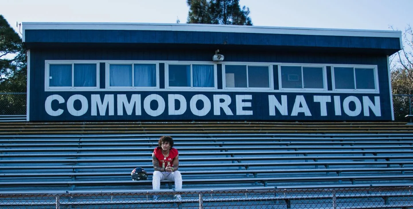 A person in a red football jersey sitting on football bleachers with a black helmet nearby, in front of a building with a large sign that reads "COMMOODORE NATION."