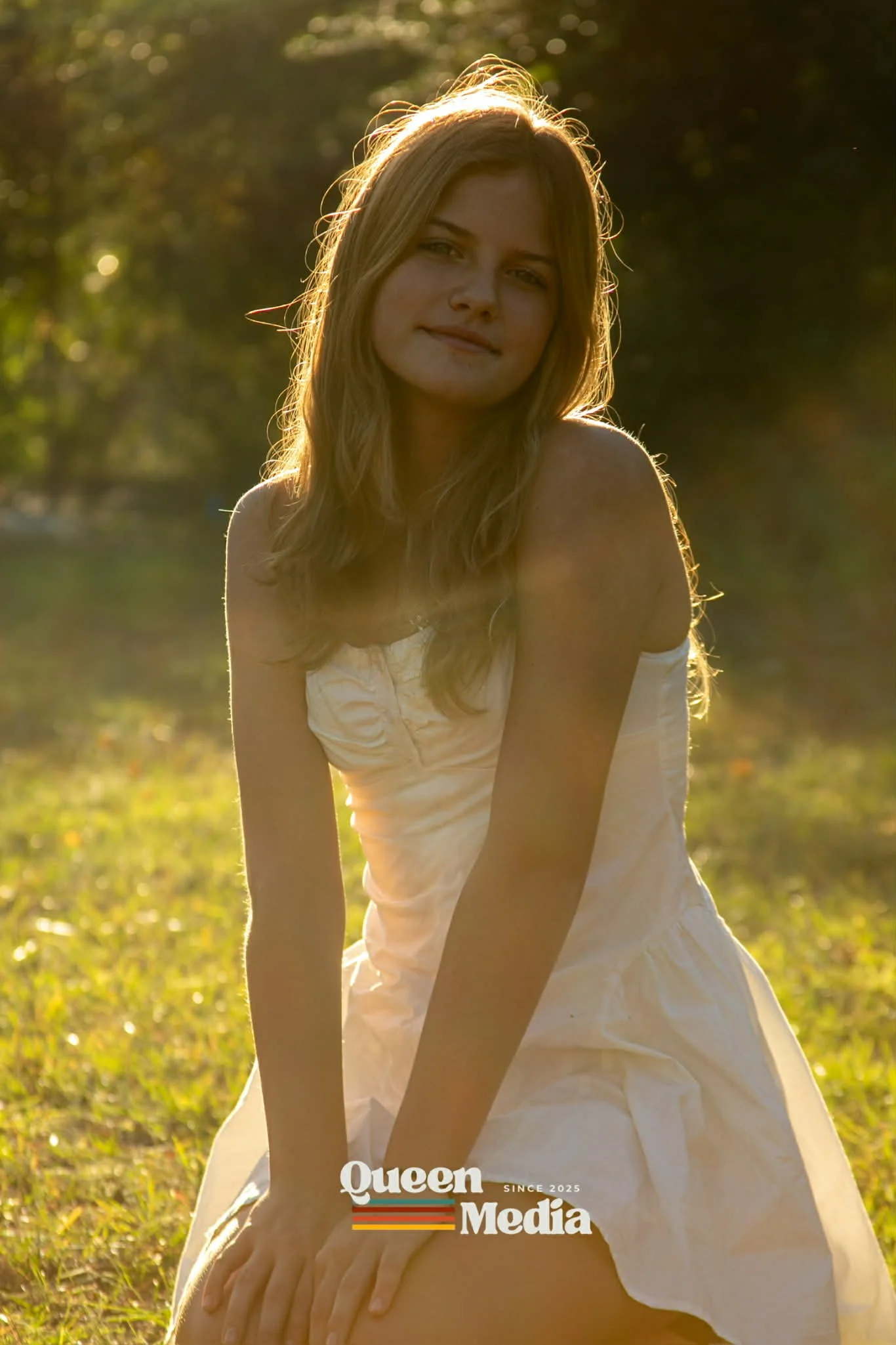 A young woman with blonde hair in a white dress, kneeling outdoors in sunlight, with a blurry green background, smiling gently at the camera.