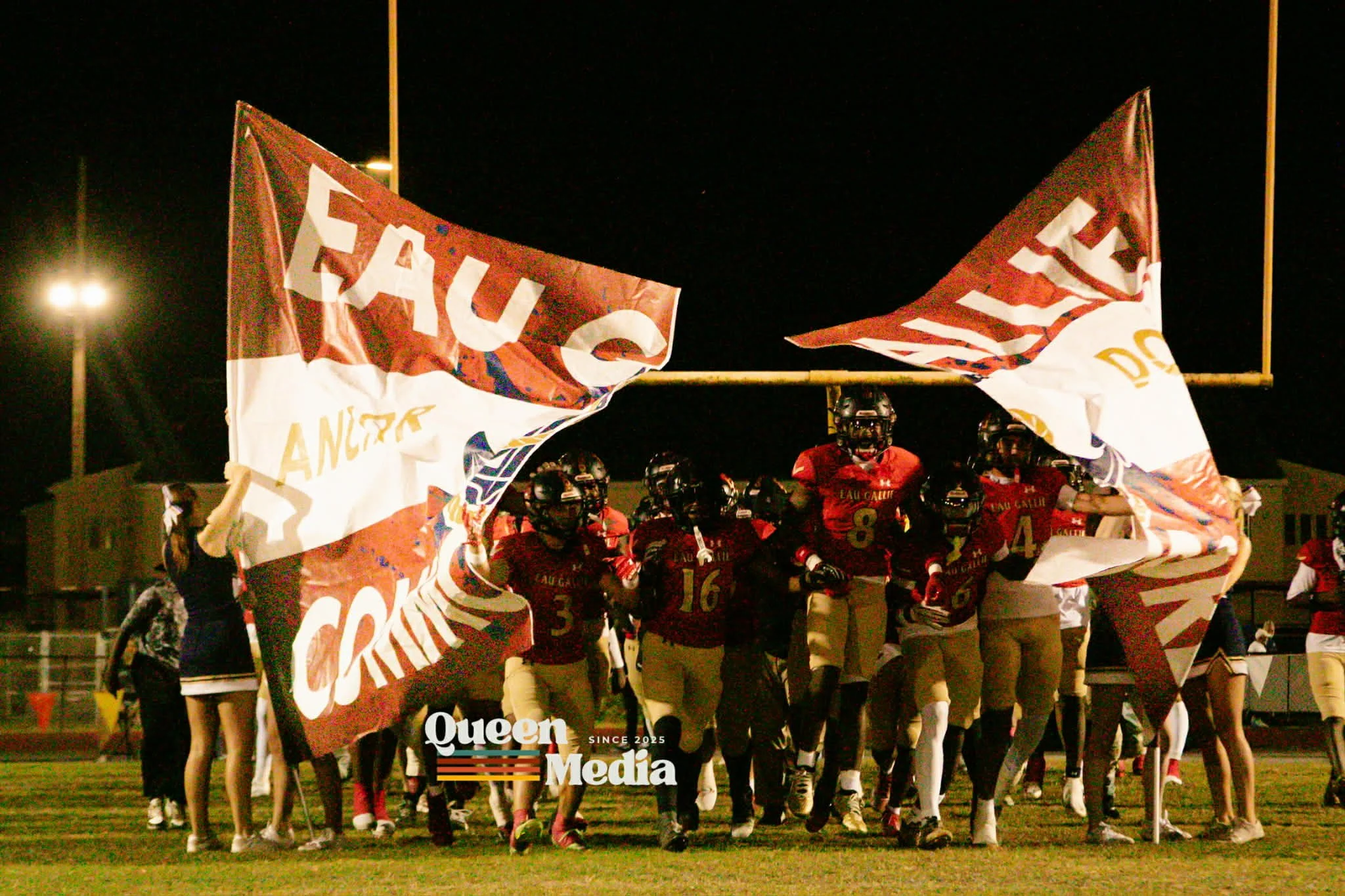 High school football team in red jerseys and tan pants running onto the field through a large banner at night, with players holding the banner. The banner displays the words 'FAU' and 'CRAWL' and possibly other words. The team is under a football goalpost, and stadium lights are visible in the background.