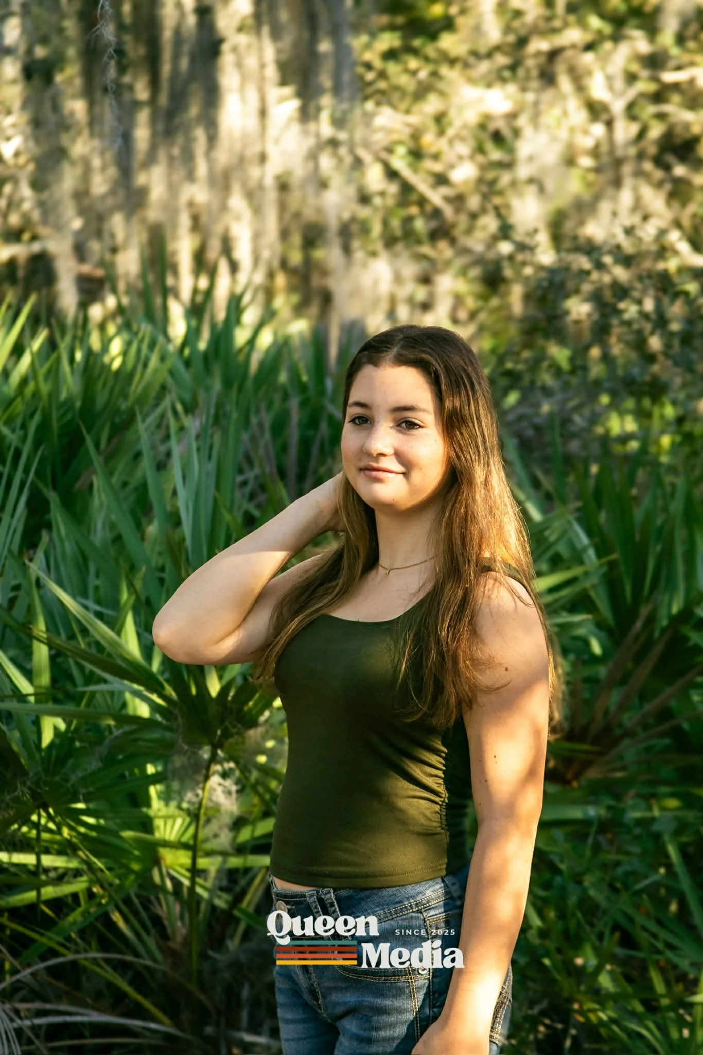 A young woman with long brown hair stands outdoors in a lush green setting with tall grasses and trees in the background. She is wearing a sleeveless black top and jeans, and has a gentle smile while touching her hair.