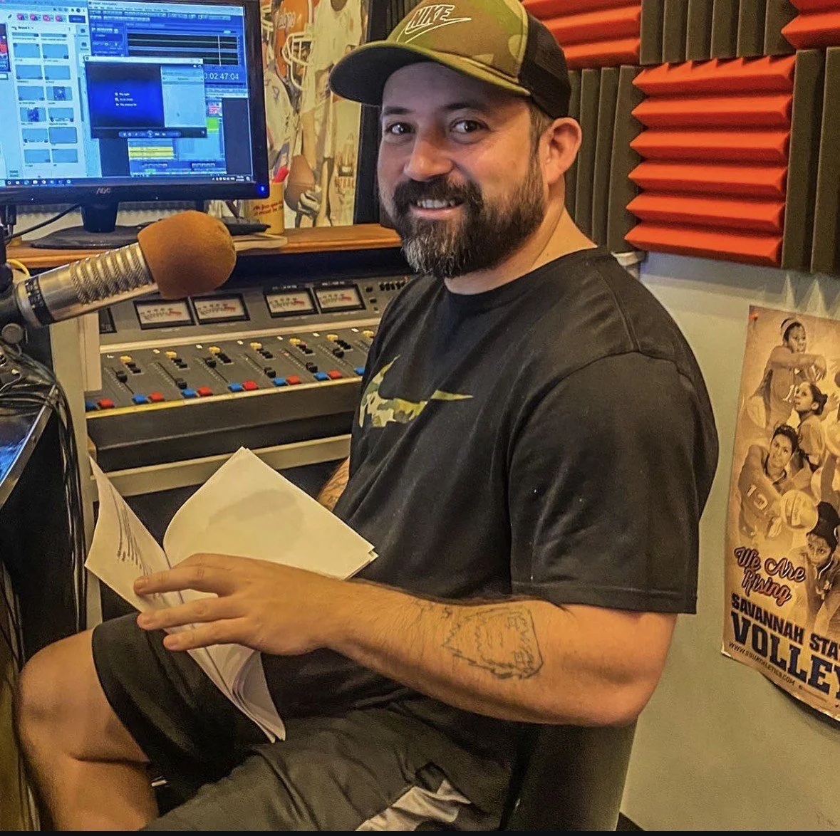 A man with a beard and a camouflage baseball cap sitting in a radio station studio, holding a script, with a microphone and sound equipment in front of him, and a computer monitor in the background.