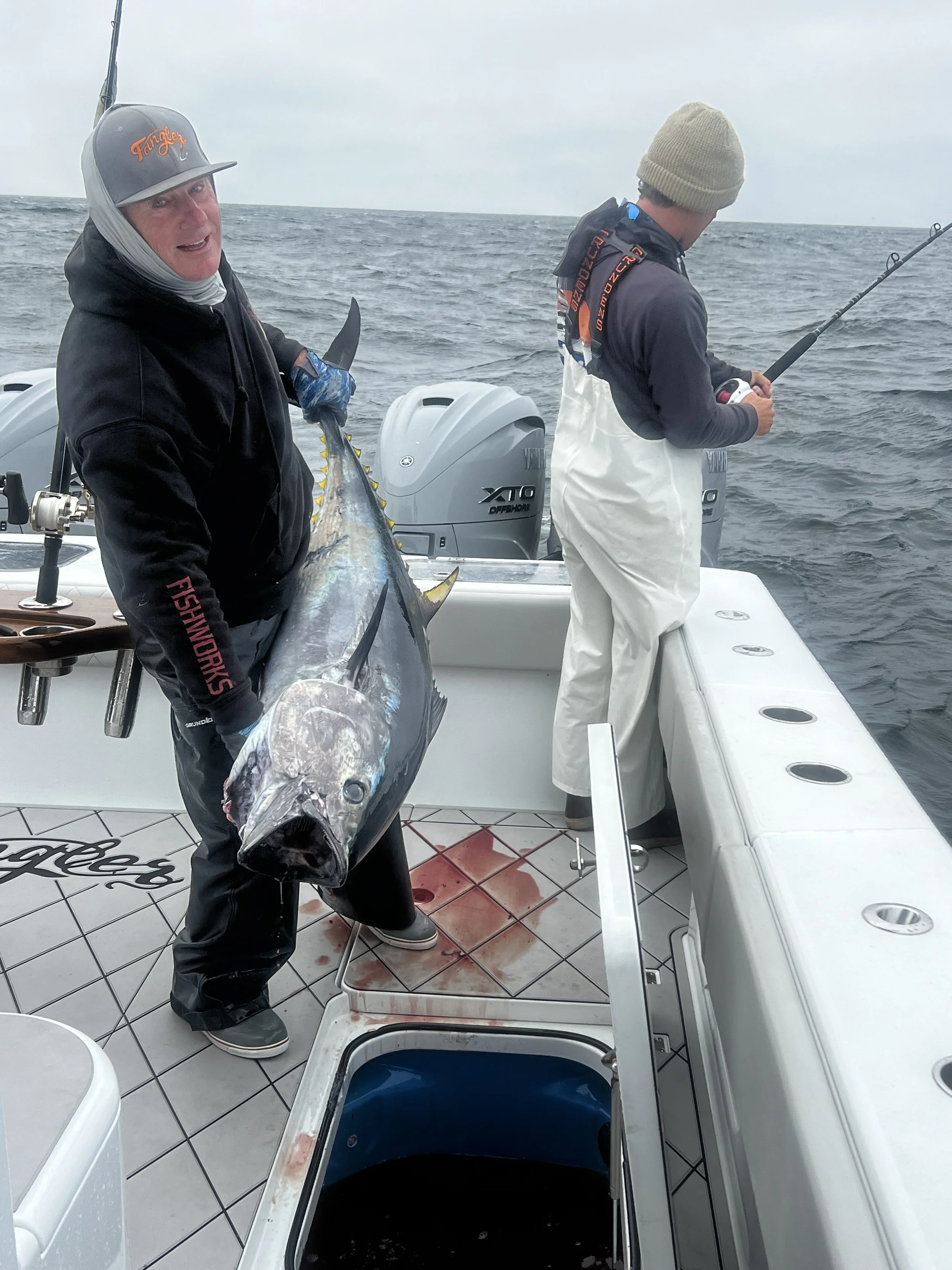 Two men on a boat in the ocean, one holding a large bluefin tuna while the other one fights another bluefin.  