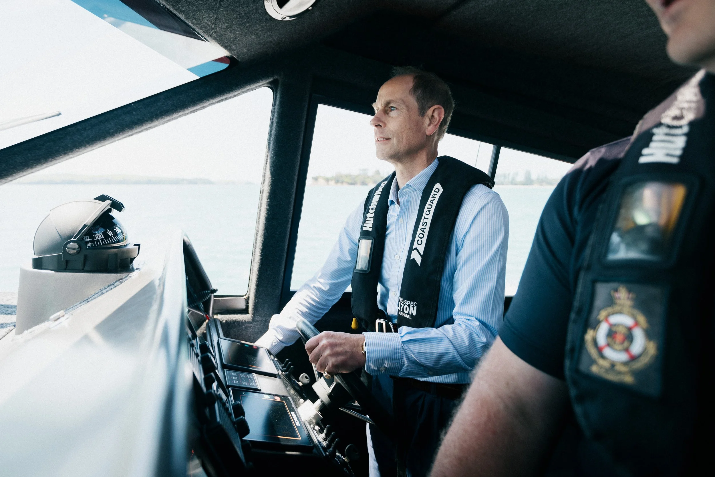 Prince Edward sitting at the controls of a coastguard boat with another person partially visible, heading back from visiting the Spirit of Adventure in Auckland. The scene is set outdoors on a body of water with a distant shoreline.