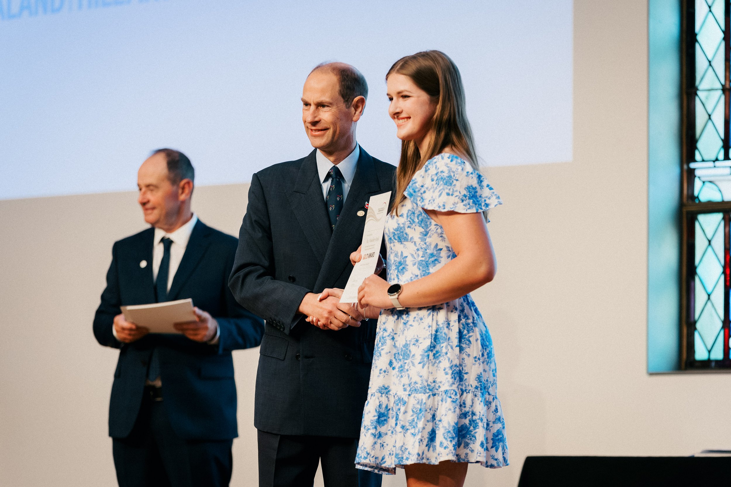 A woman in a blue floral dress receiving an award, presented by a man in a suit. Another man in a suit is standing to the side holding a folder.