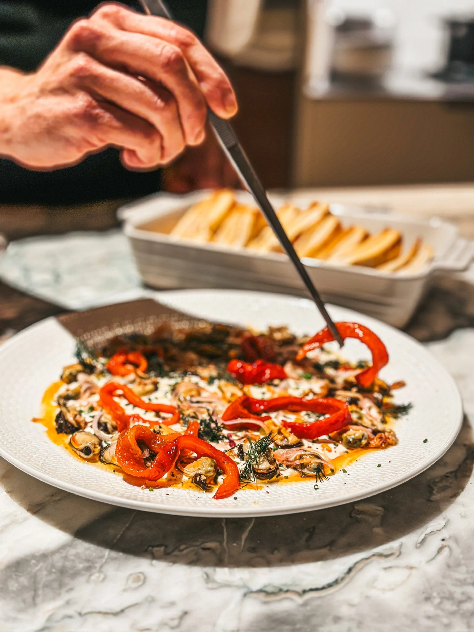 Person drizzling sauce over a gourmet seafood dish with mussels, red peppers, herbs, and cream sauce, served with fries.