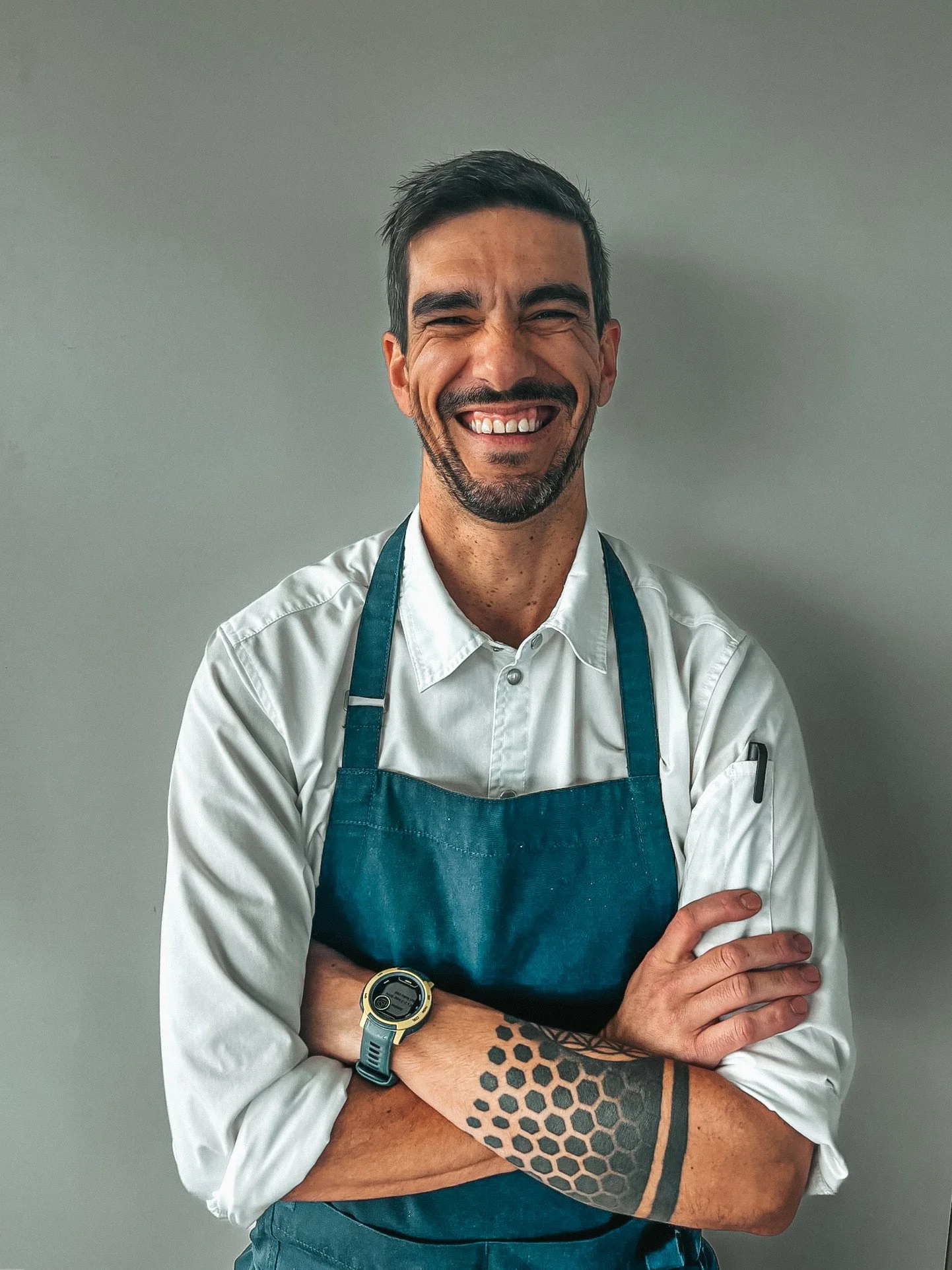 Smiling bearded man in a white shirt and teal apron with crossed arms