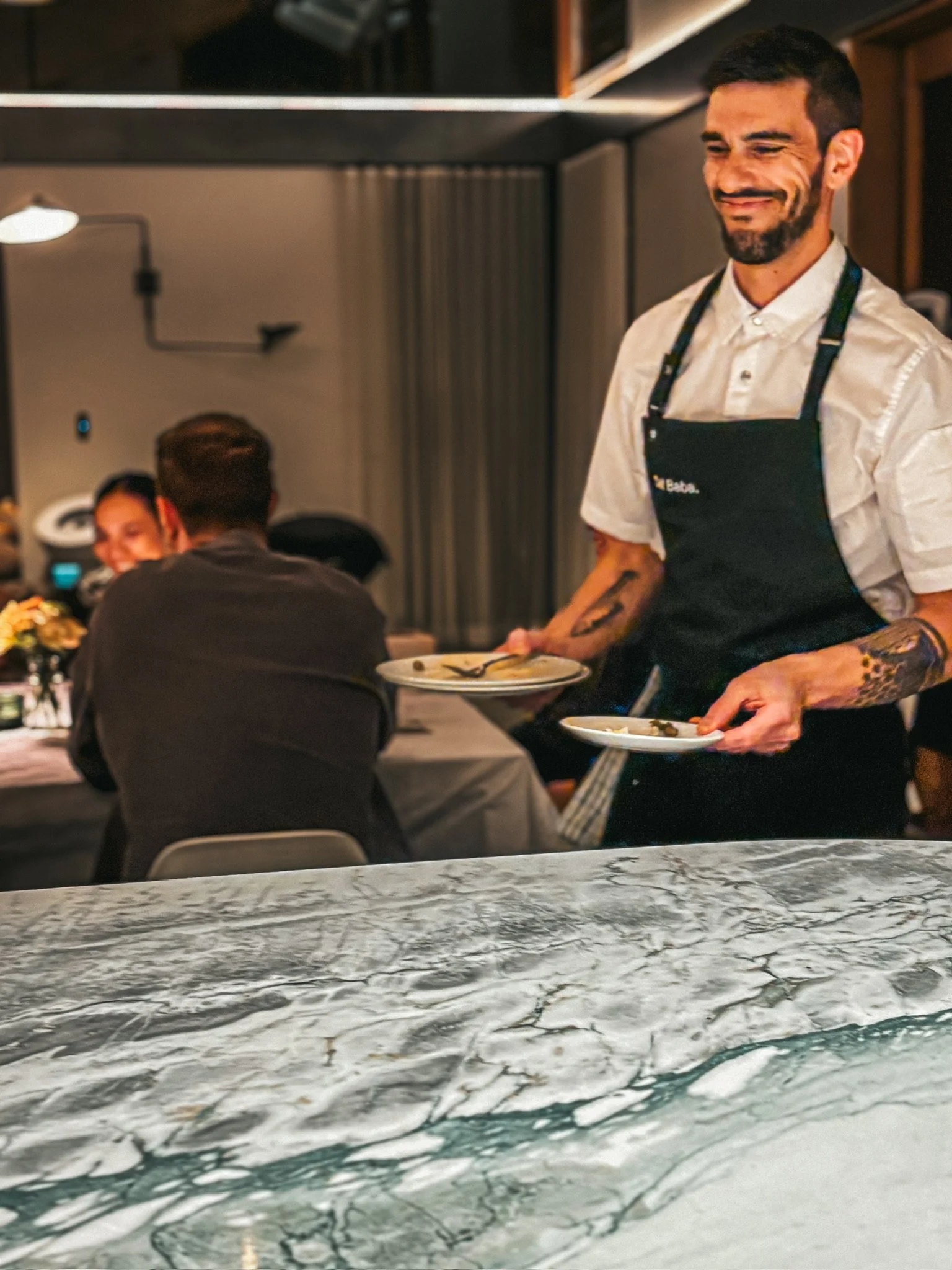 Smiling restaurant server in a black apron carrying plated dishes to diners.