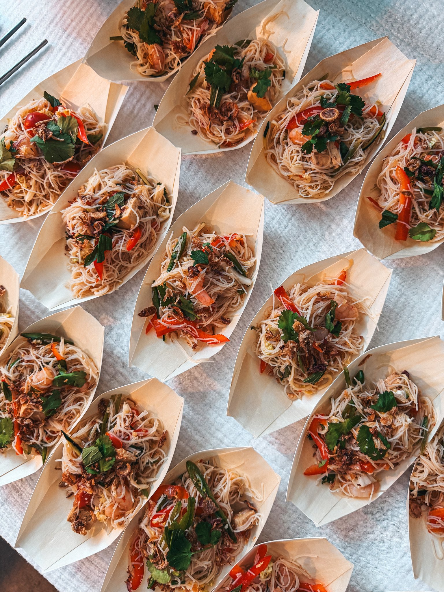 Overhead view of numerous takeaway boxes filled with rice noodle salads, shrimp, vegetables, cilantro, and crispy toppings