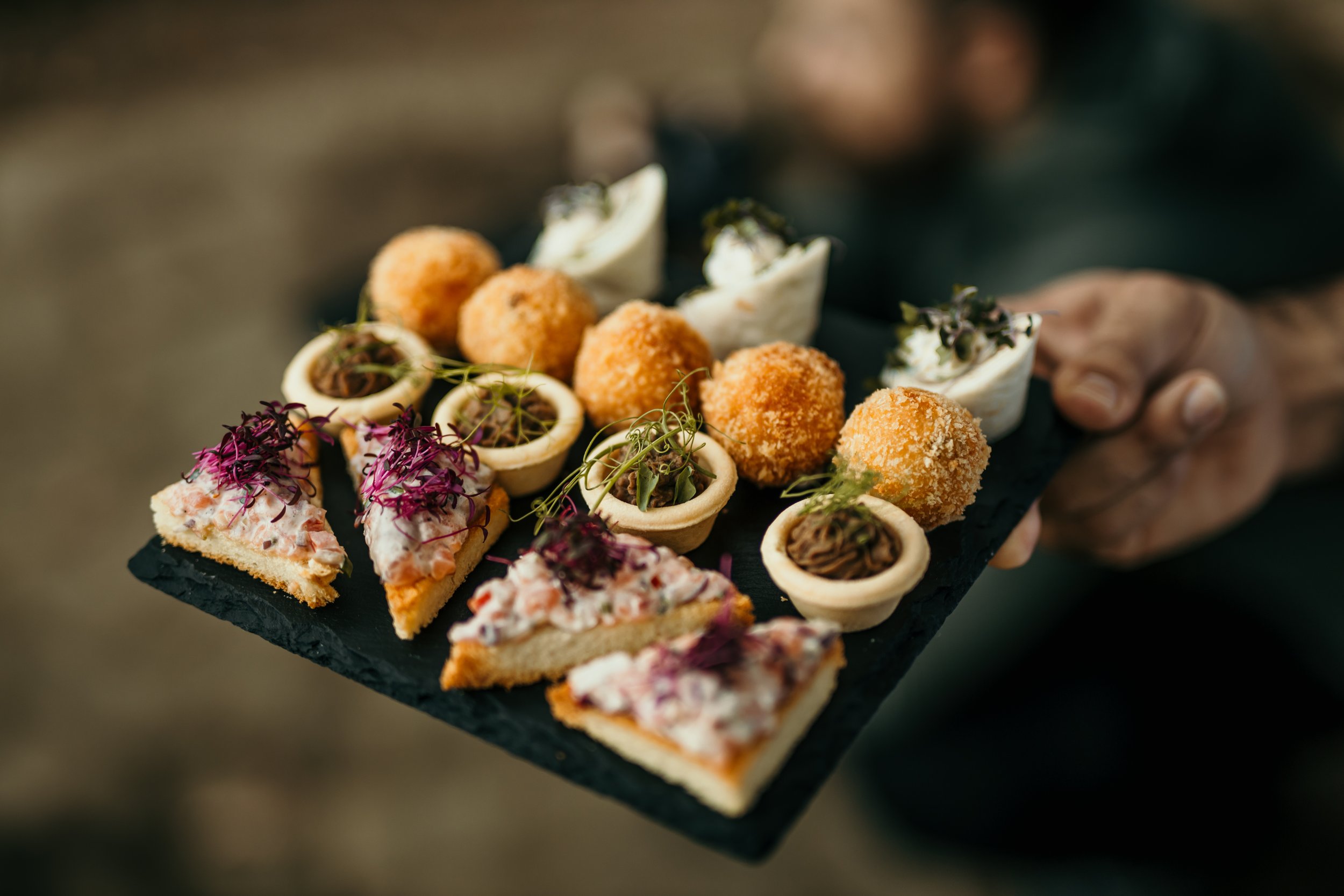 Assorted gourmet hors d'oeuvres on a black slate serving tray, including croquettes, savory tartlets, and garnished canapés