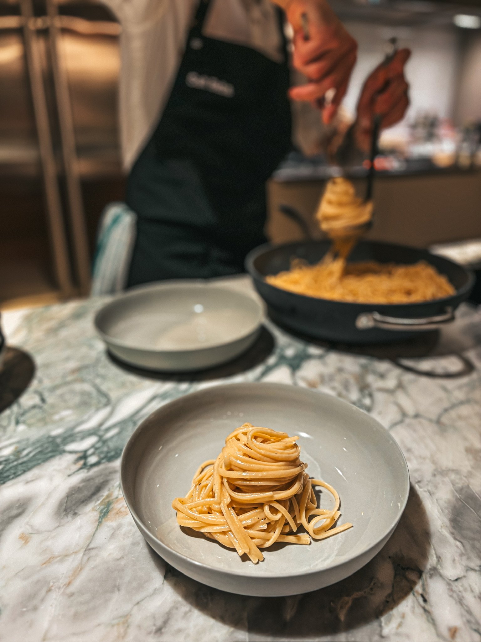 Chef plating spaghetti in a pasta restaurant kitchen, with a plated serving in the foreground