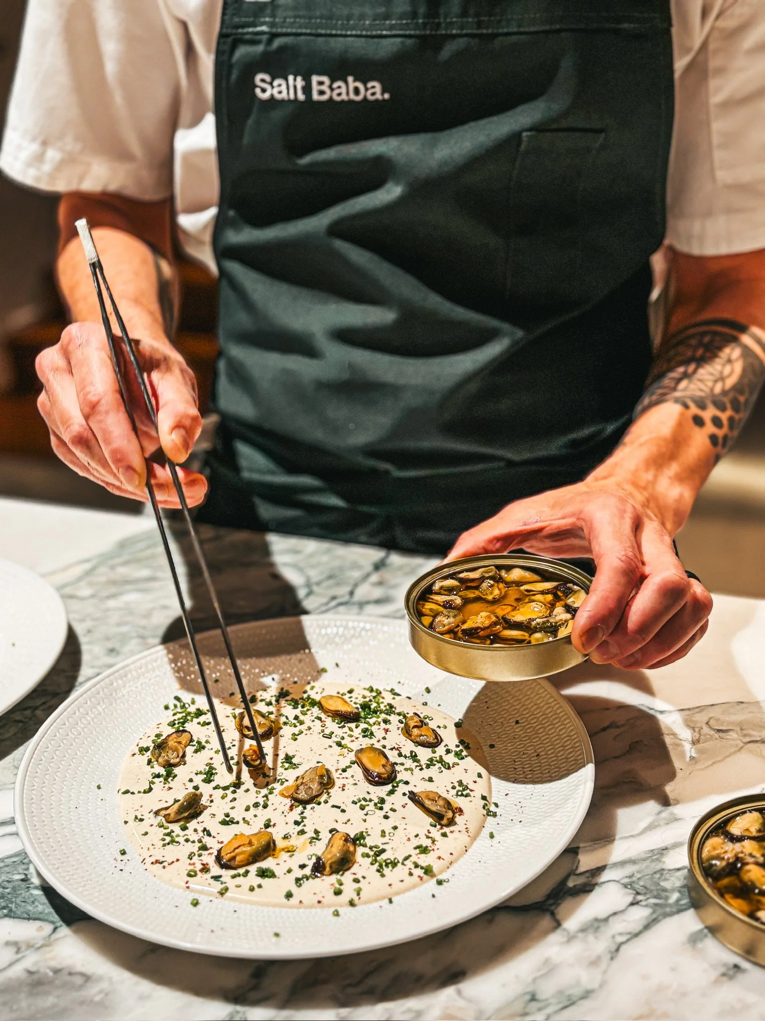 Chef serving mussels from a tin onto a creamy sauce plate with chopsticks.