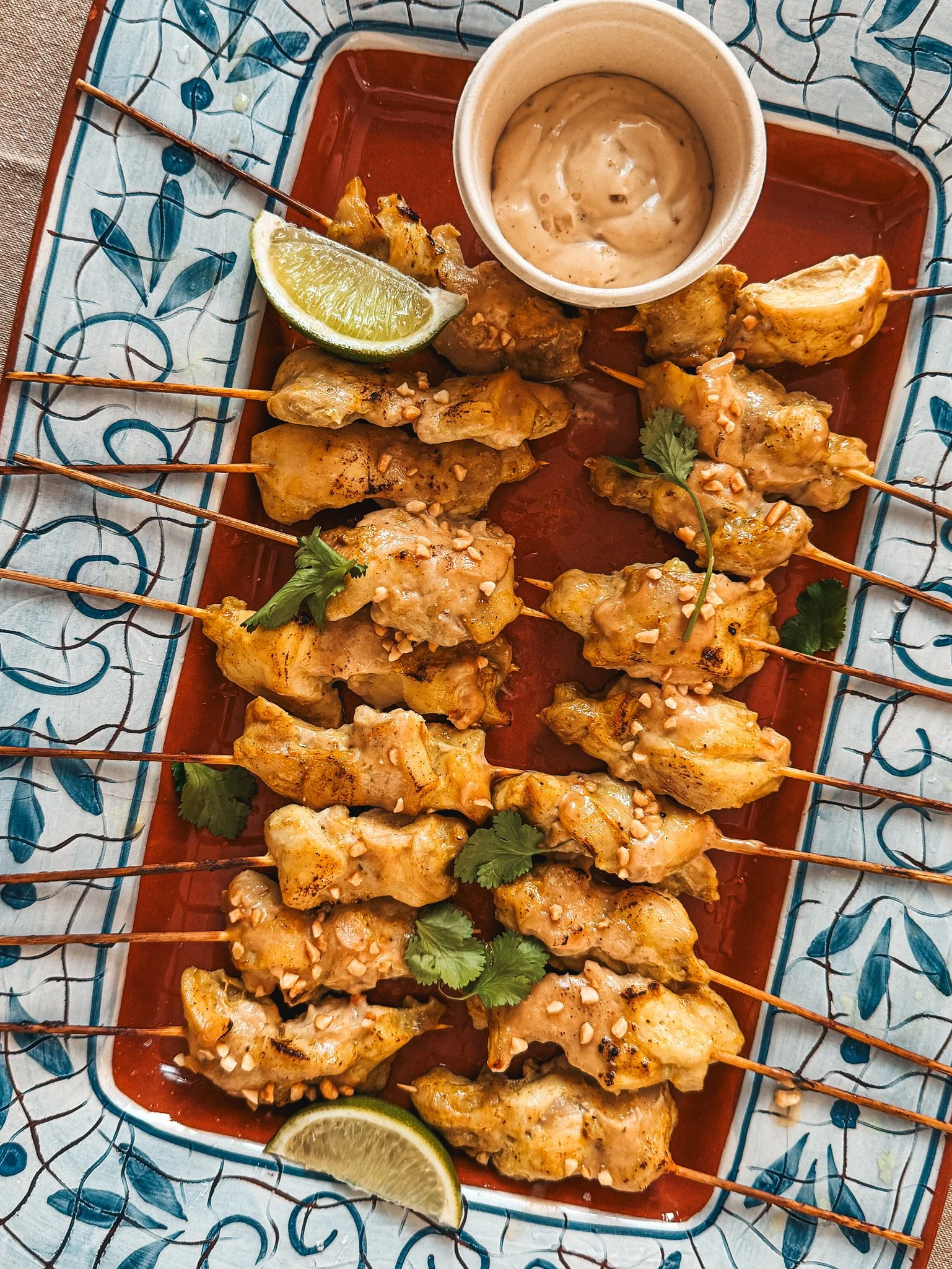 Grilled chicken skewers with peanut sauce, lime wedges, chopped peanuts, and cilantro on a decorative platter