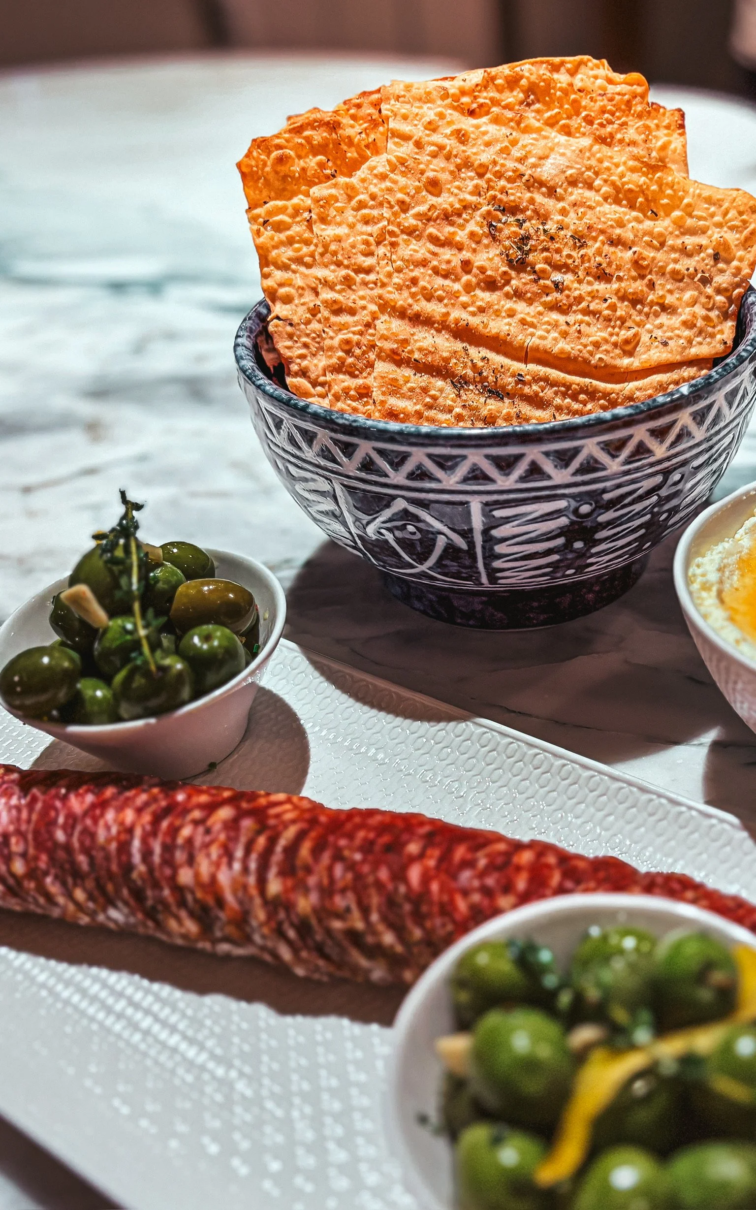 A bowl of crispy orange crackers, small bowls of green olives, and a long cured sausage on a white tray.