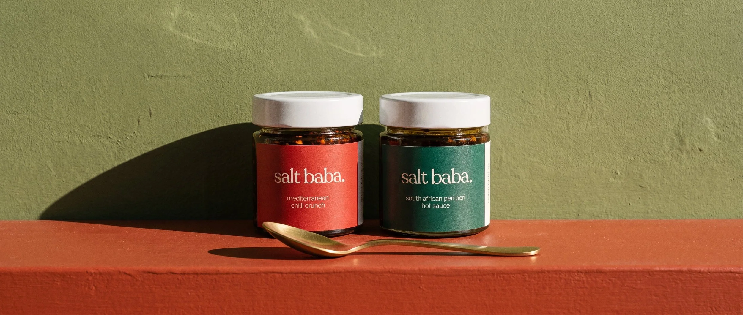 Two Salt Baba spice jars labeled Mediterranean chilli crunch and South African peri peri hot sauce beside gold spoons.