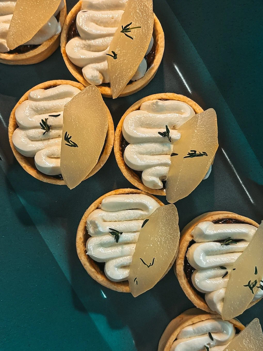Assorted fruit tartlets topped with piped white cream, translucent fruit slices, and small herb garnishes on a dark teal tray