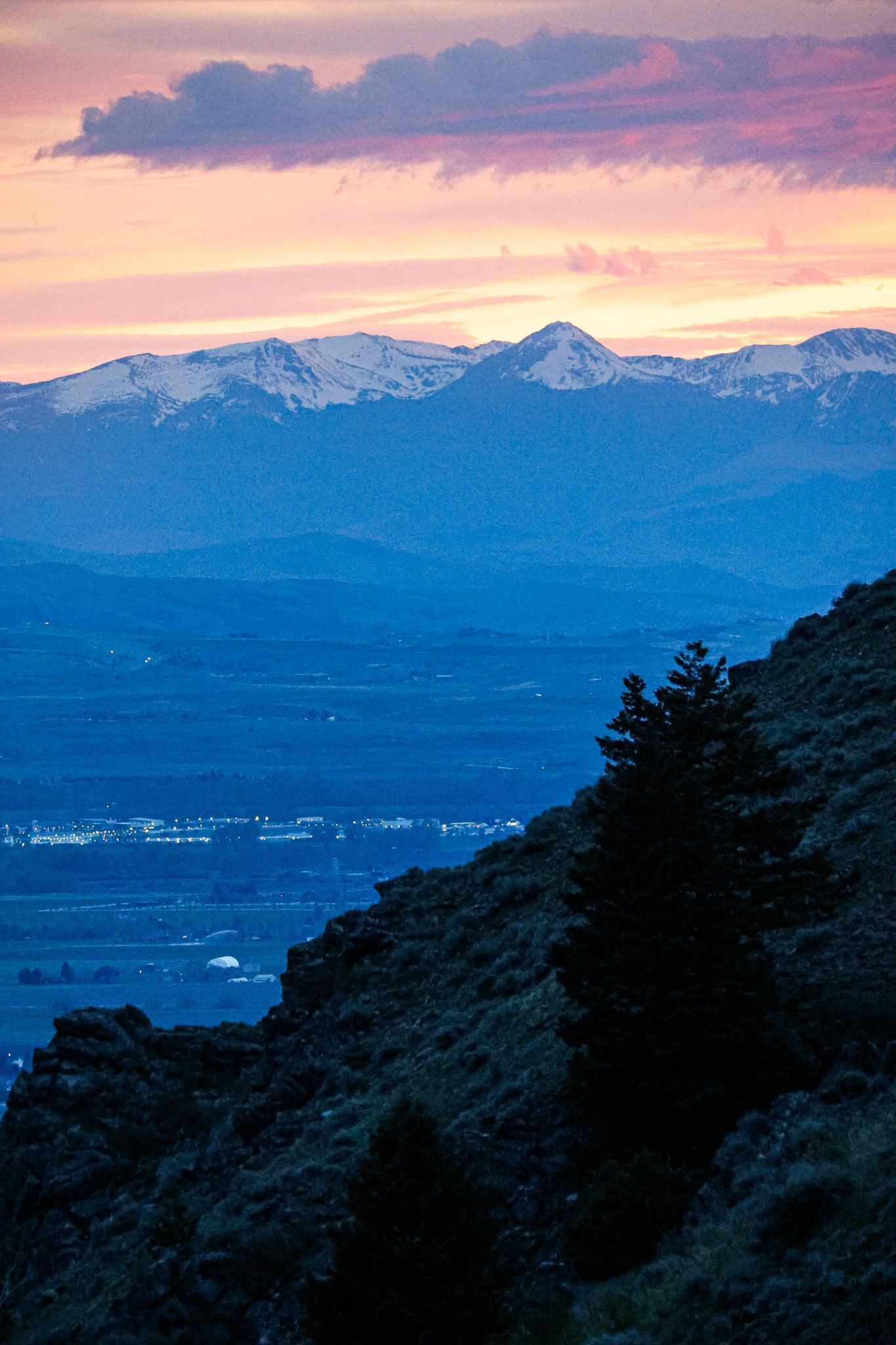 Scenic view of snow-capped mountains at sunset with purple and pink sky, dark forest in the foreground, and faint city lights below.