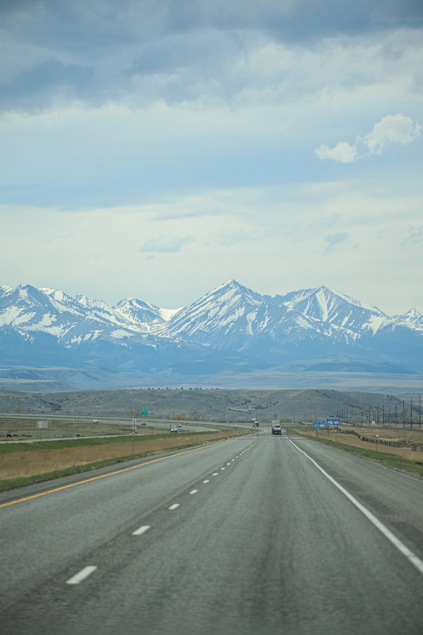 View of a highway leading towards snow-capped mountains in the distance under a cloudy sky.