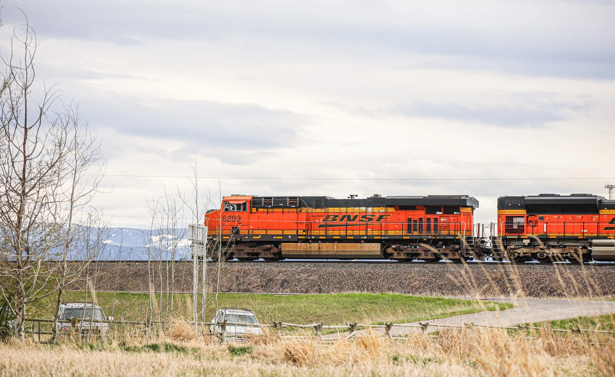 A BNSF freight train engine moving on tracks through a rural area with bare trees and distant mountains under a cloudy sky.