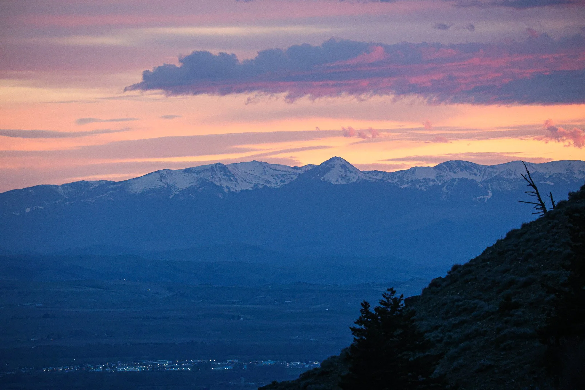A scenic landscape featuring snow-capped mountains at sunset with a sky painted in shades of pink, purple, and orange, and a dark silhouette of trees in the foreground.