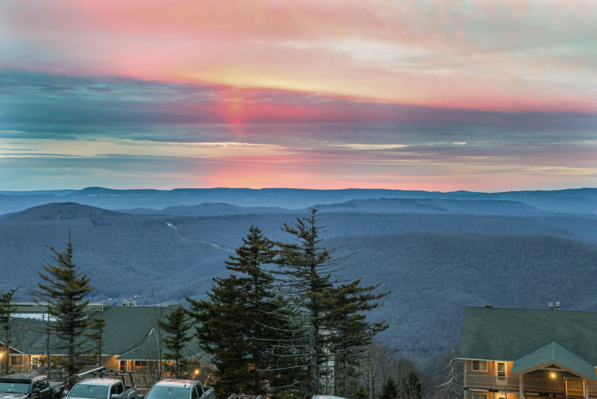 A scenic view of mountains and a colorful sunset with pink, purple, and blue sky overhead. There are trees in the foreground, some buildings with rooftops, and a parking lot with cars at the bottom of the image.