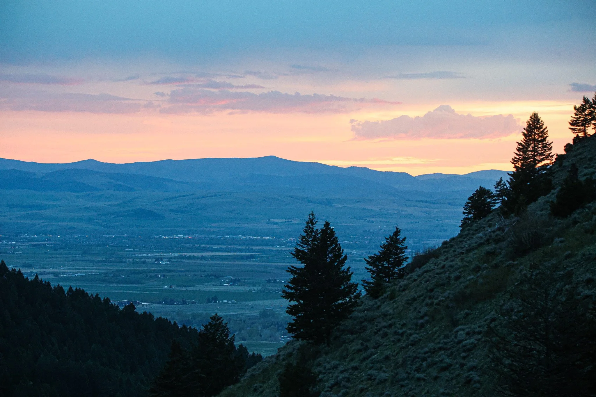 A scenic mountainous landscape at sunset with layered mountain ridges, a valley, and trees in the foreground.