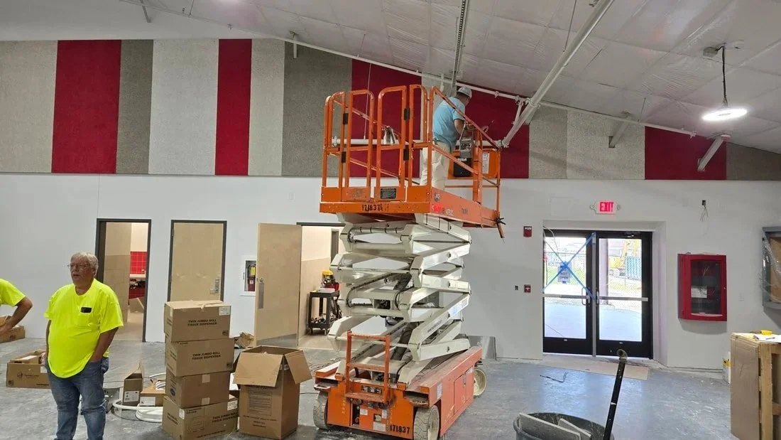 Workers installing ceiling panels using a scissor lift, with boxes and tools on the floor, in a commercial building under construction.