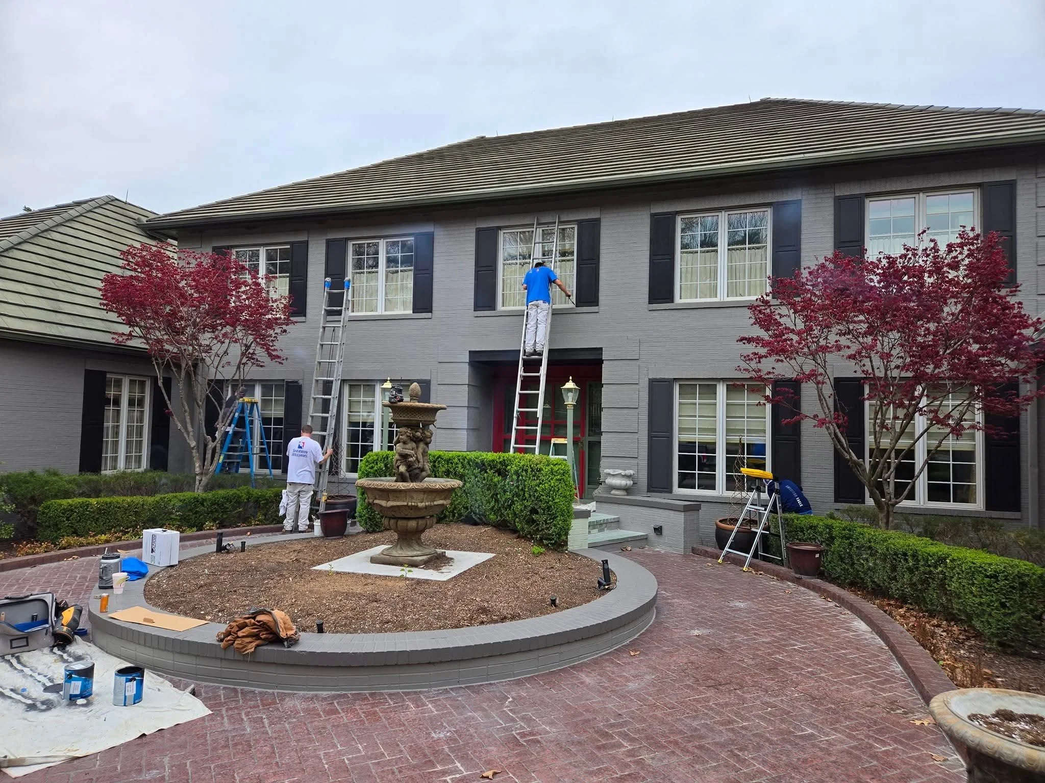 Workers painting the outside of a gray house with black shutters, using ladders, with a landscaped front yard featuring trees, a fountain, and a brick walkway.