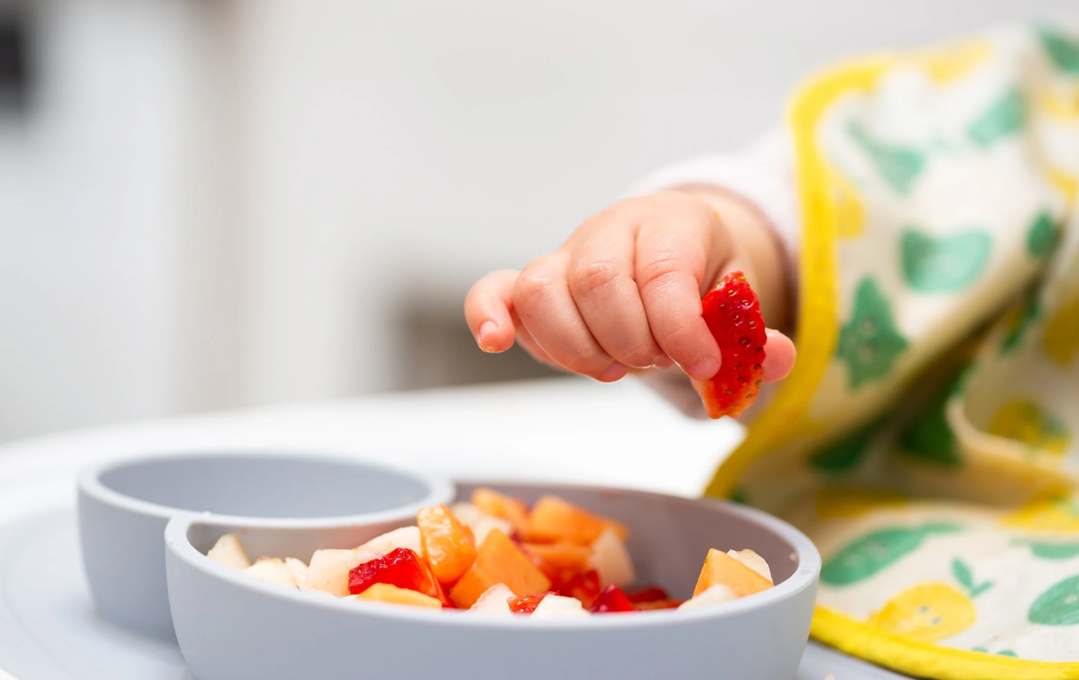 A child's hand holding a piece of strawberry over a bowl of chopped fruit.