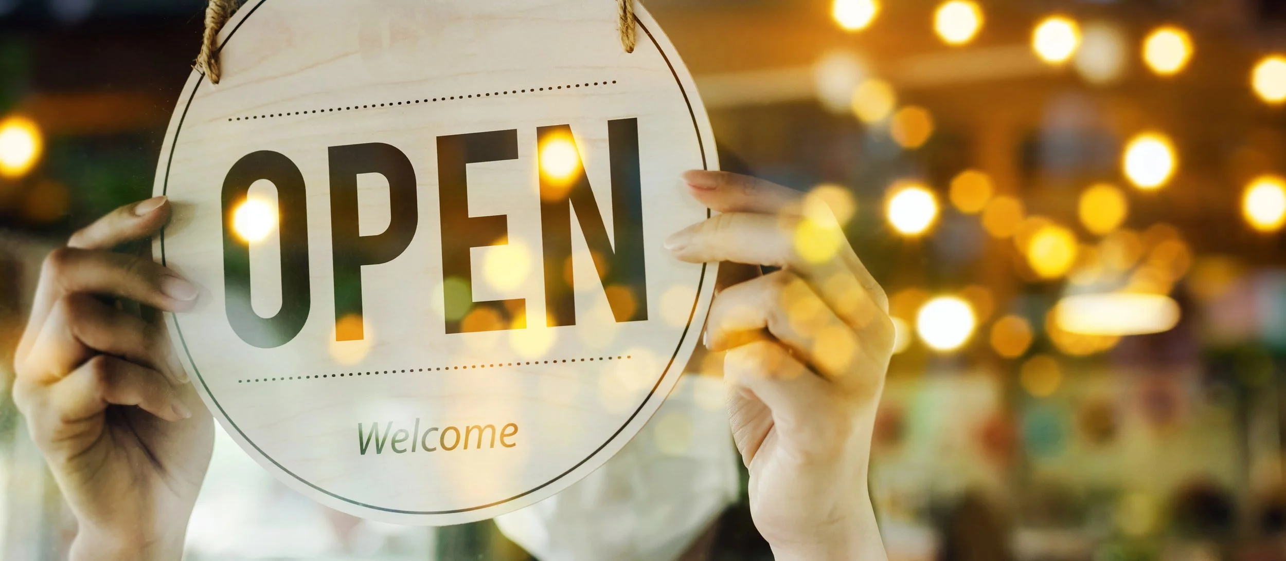 Hands holding a wooden sign that says 'OPEN' with a welcoming message underneath, illuminated with warm bokeh lights in the background.