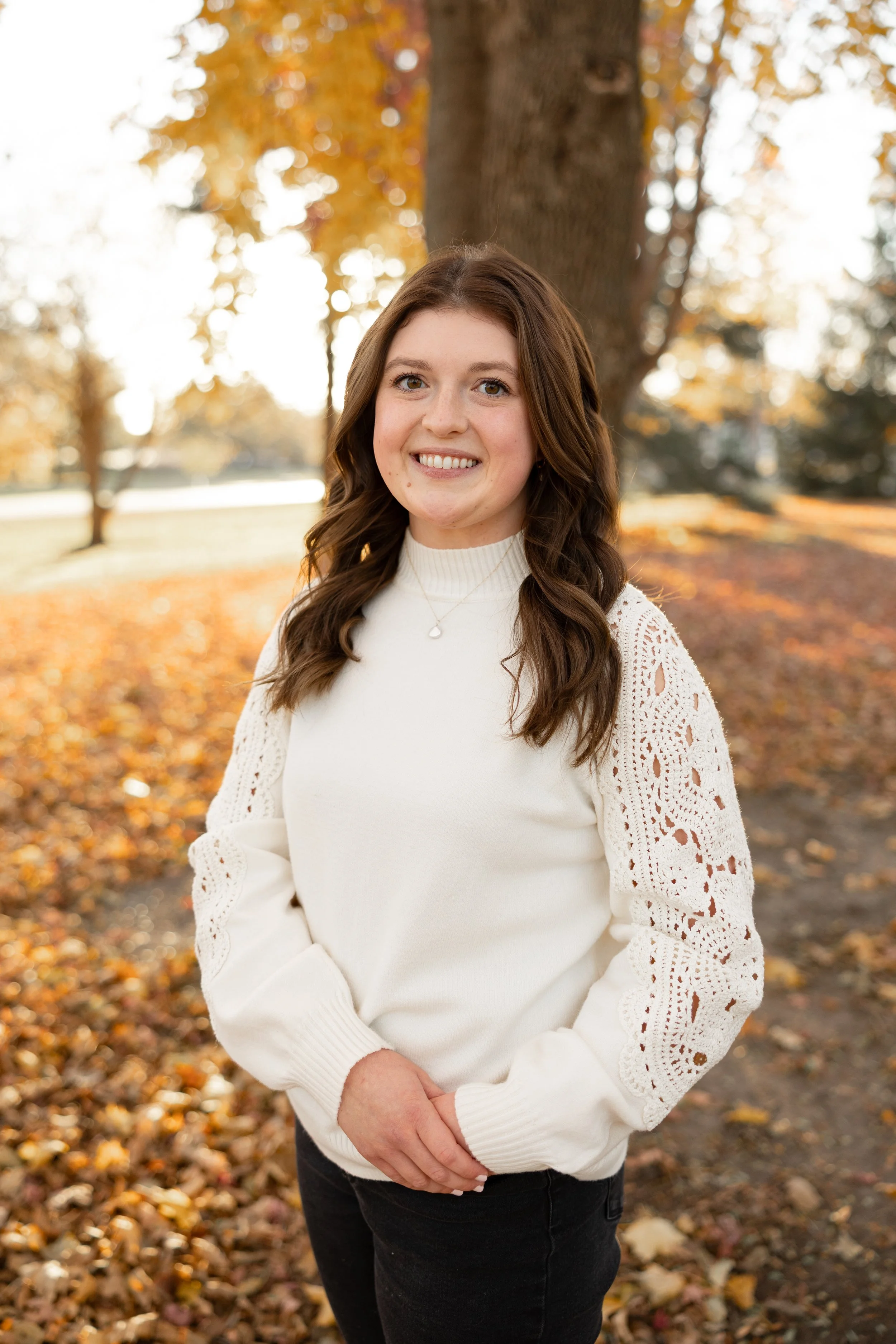 A woman with long brown wavy hair smiling outdoors in autumn, wearing a white sweater with lace details on the sleeves, standing in front of a large tree with fall foliage.