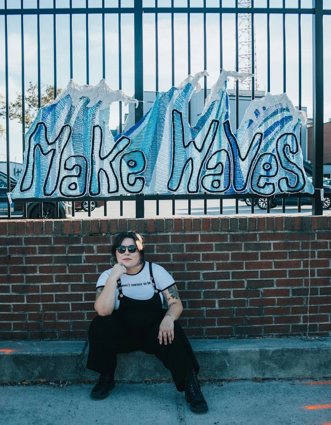 A woman sitting on a sidewalk in front of a brick wall with a sign that reads 'Make Waves' with wave illustrations, wearing sunglasses, black overalls, a white t-shirt, and black boots.